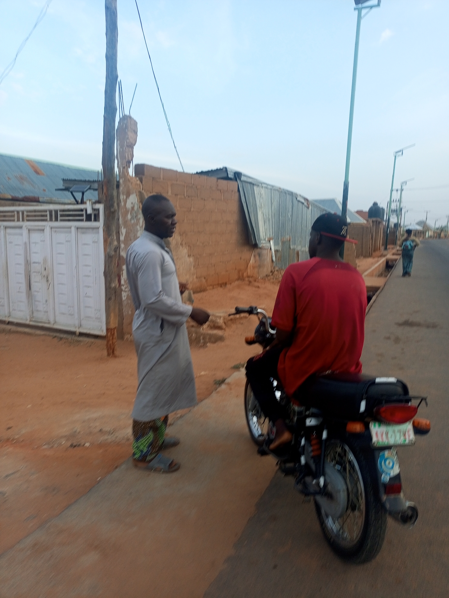 Two men are standing on a street next to a motorcycle. One man is dressed in a long gray garment with patterned pants and sandals. The other man is sitting on the motorcycle wearing a red shirt and a black cap with a red brim. A woman can be seen walking in the distance. The street is in a residential area with buildings and fences visible in the background. There are streetlights along the road.