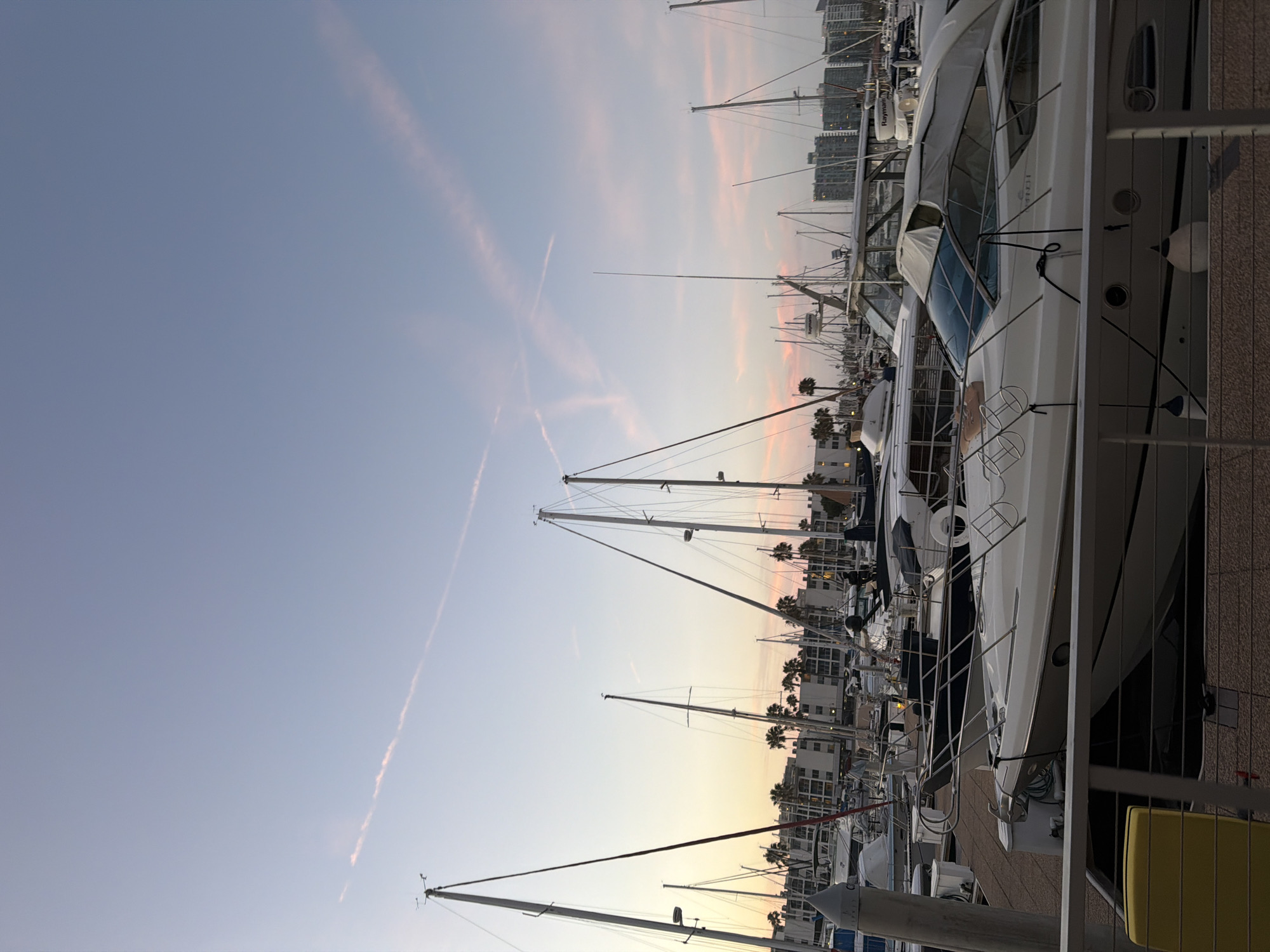 The image shows a marina with numerous sailboats and yachts docked. The sky is a gradient of soft blue and pink, suggesting either dawn or dusk. Contrails are visible in the sky, indicating recent aircraft activity. Palm trees are scattered throughout the background, and buildings with a coastal architectural style line the waterfront. In the foreground, a portion of a white yacht with a canvas cover is visible, along with a yellow bollard and a metal railing. The scene is calm and serene, typical of a harbor at the end or beginning of the day. The specific location is Marina del Rey in Los Angeles, California, United States of America, known for its large boat harbor and waterfront residences. There are no people clearly visible in the image, and the focus is on the boats and the sky.