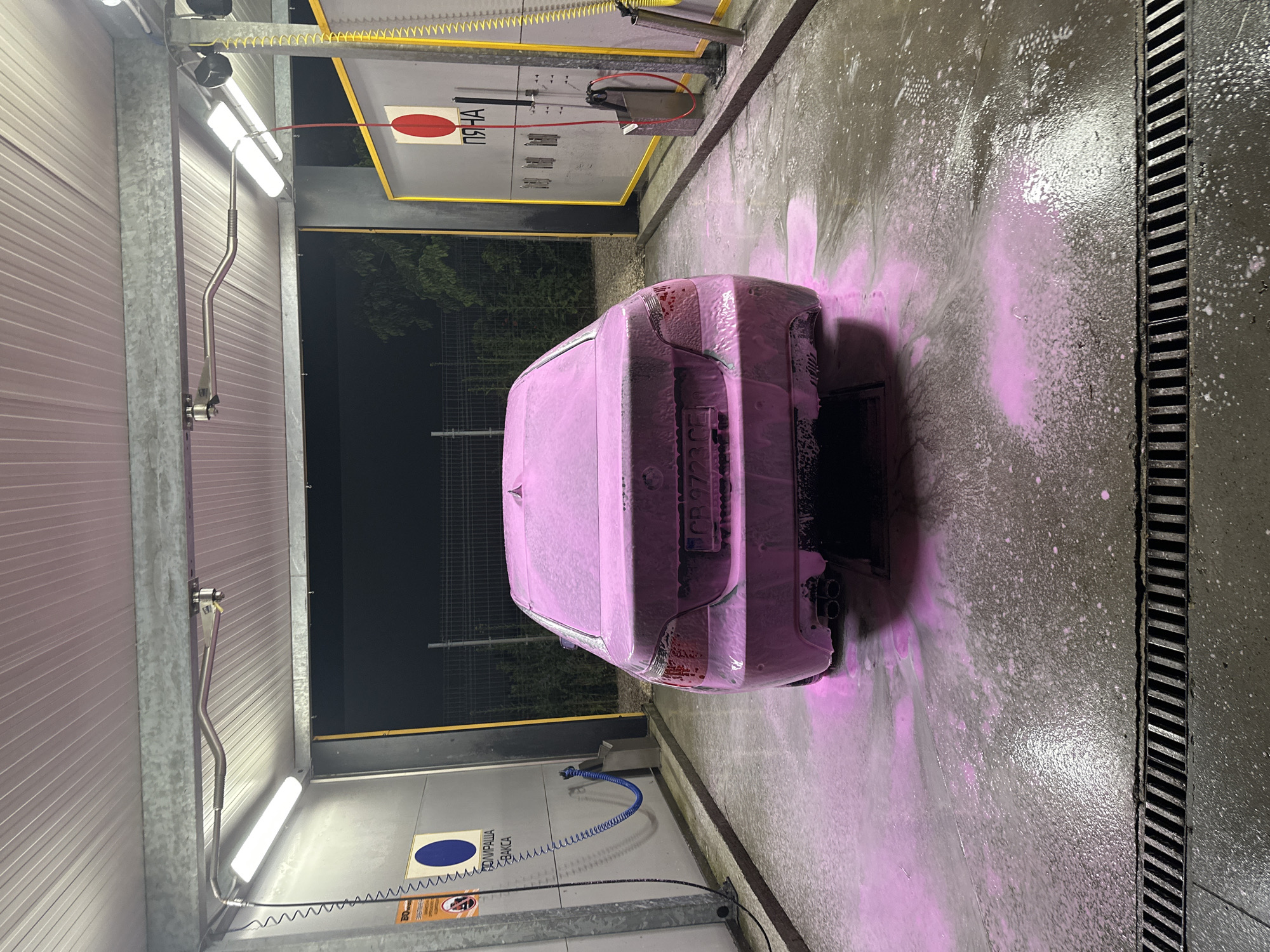 The image shows a car covered in pink foam at a car wash. The car is facing away from the camera. In the background, there's car wash equipment on the wall and overhead lighting. The location is Plovdiv, Bulgaria, as indicated by the license plate on the car. The ground is wet and foamy, leading to a drain. The environment is enclosed.