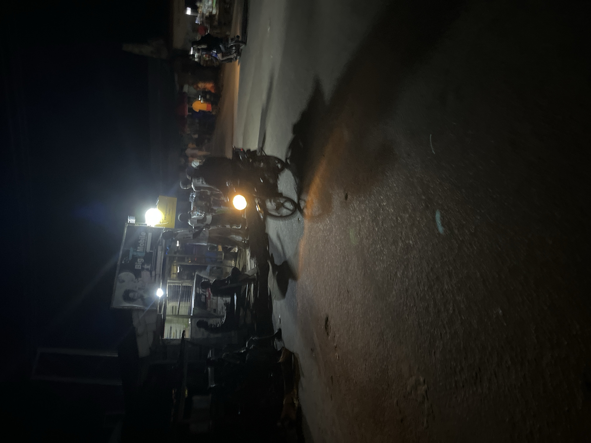 In the city of Unknown, at night, a motorcycle with its headlight on is the primary subject, casting a bright yellow glow on the asphalt road. The motorcycle is in motion, indicated by the blurred wheel and the distinct shadow it casts. Several people are visible in the background, some standing and some seated near a building with a "Big Mobile" sign. The scene appears to be a street in a town or city, with some structures and other vehicles present in the distance. The overall atmosphere is dimly lit, characteristic of a nighttime urban environment.