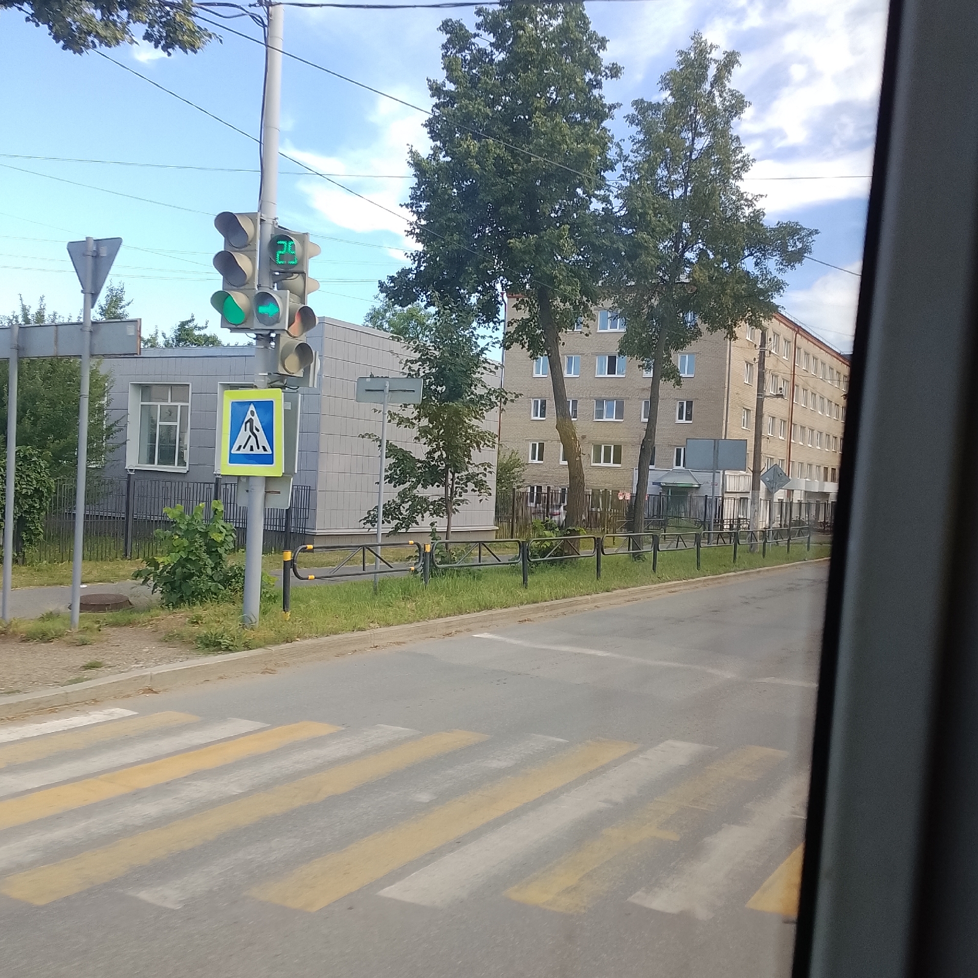 The image captures a street scene in Chaikovsky, Russia, on a sunny day with a blue sky and scattered clouds. In the foreground, a pedestrian crossing with yellow and white stripes is visible on the asphalt road. To the left of the crossing, a traffic light displays a green light with an arrow pointing left, and a countdown timer showing "29". A pedestrian crossing sign, a blue square with a white triangle containing a black silhouette of a person walking, is mounted on a pole near the traffic light.
In the middle ground, a modern, grey, two-story building with a regular pattern of windows stands to the left, bordered by a black metal fence and lush green bushes and trees. Behind the pedestrian crossing and the initial building, a taller, light-brown, brick apartment building with numerous windows stretches across the background. Several mature trees with dense green foliage are interspersed between the buildings. A black metal fence runs along the edge of the sidewalk in front of the apartment building. The image appears to be taken from inside a vehicle, with the right edge of the frame showing a dark, vertical element, likely part of the vehicle's interior.