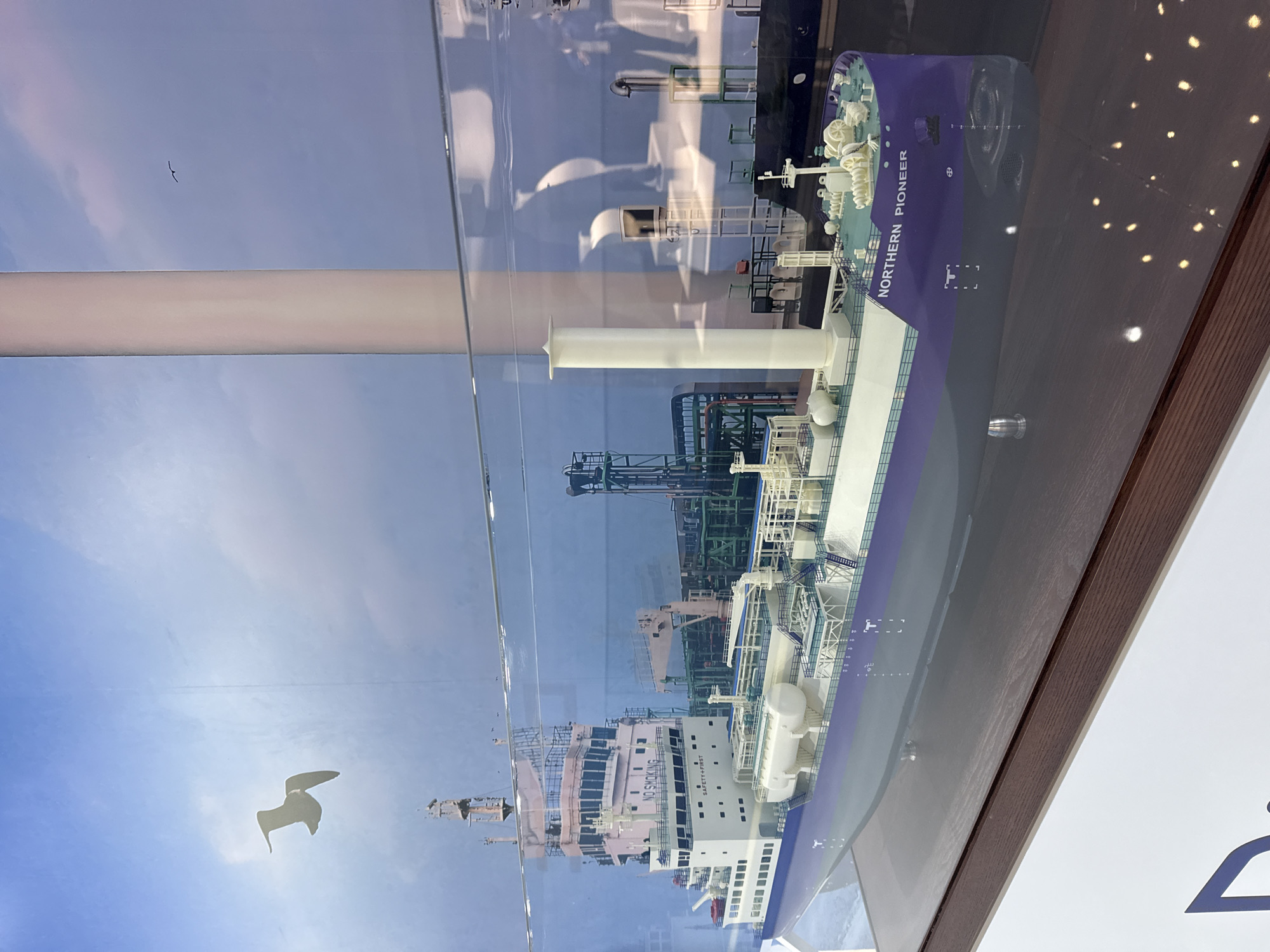 A detailed model of a ship, identified as the "Northern Pioneer," is displayed behind glass, likely in a museum or exhibition. The ship model is predominantly white and purple, featuring intricate details of its deck, superstructure, and cargo tanks. The backdrop behind the model appears to be a scenic sky with clouds and a silhouette of a large, white cylindrical structure, possibly a smokestack or a tower. Two seagulls are depicted in the sky, one in the foreground flying left and another smaller one in the distance. The lighting in the scene creates reflections on the glass, obscuring some of the background details. There are no people visible in the immediate foreground or interacting with the model. The image does not provide any information about its specific location within Bangkok, Thailand.