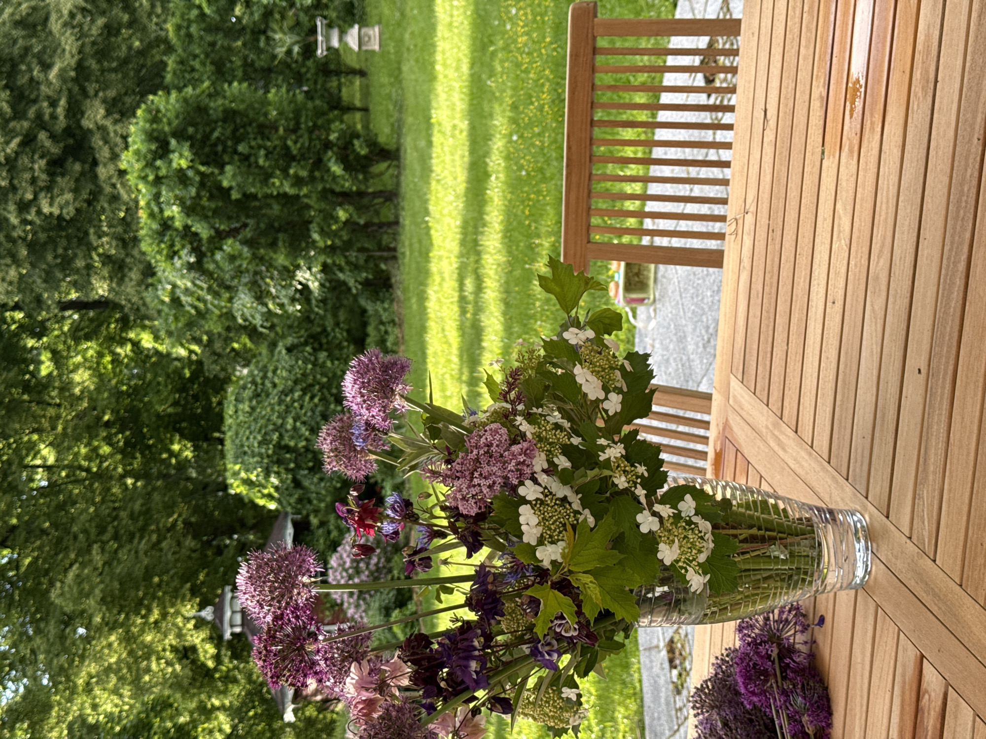 A close-up shot depicts a vase of flowers placed on a wooden table, likely outdoors given the backdrop of greenery. The vase holds a mix of purple and white flowers with green foliage. The table is made of light brown wooden slats. Behind the table, there is a green lawn and trees in the background. In the far background, there is a small structure reminiscent of a birdhouse on a pole. The image is well-lit with natural light, suggesting a sunny day. Without additional information, pinpointing the exact location in Sapronovo, Russia, or any human activity, is impossible.
