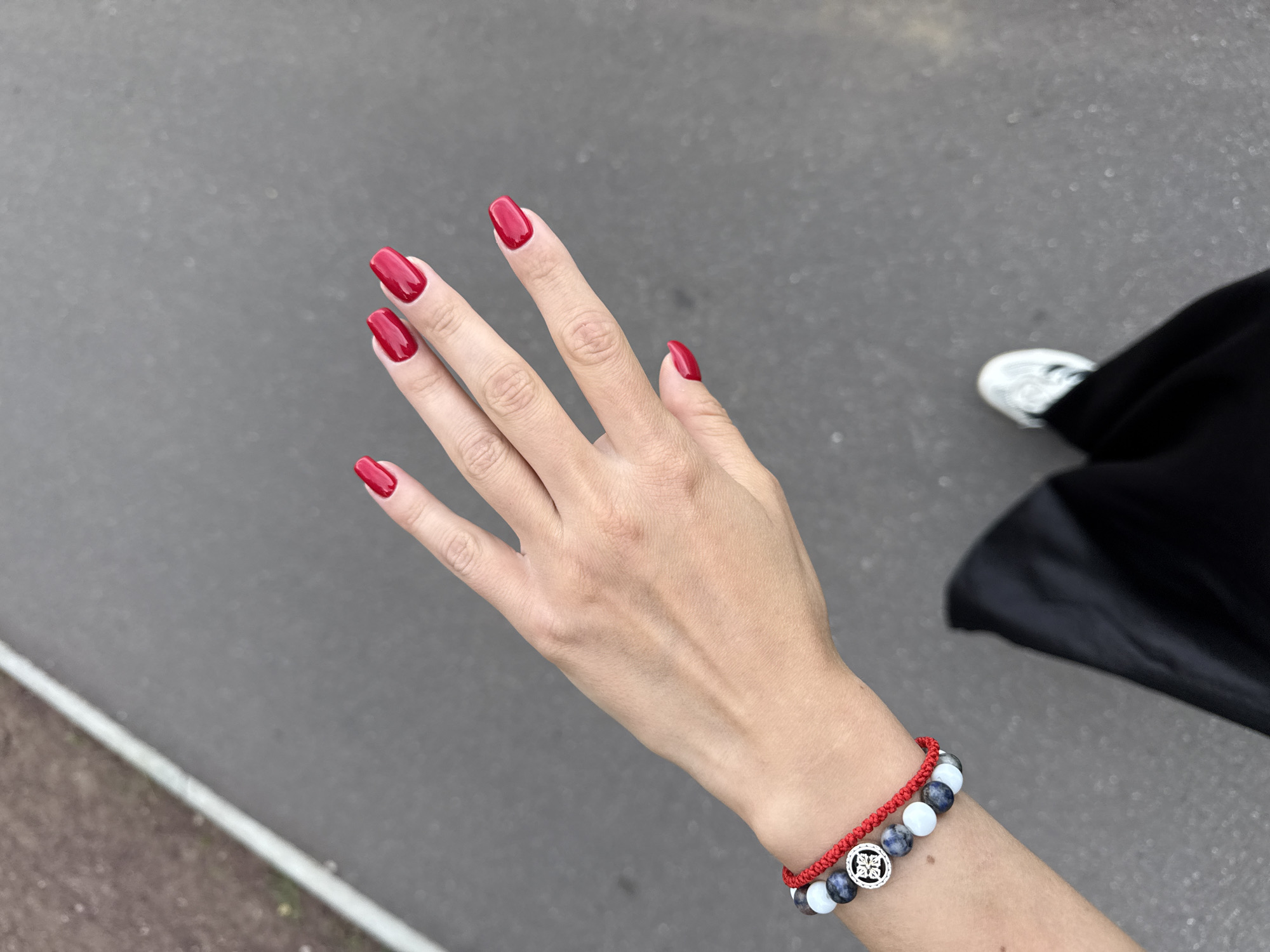 The image shows a close-up of a person's left hand and forearm, held up against a grey, asphalt-like background. The hand has bright red, polished fingernails. On the forearm, there is a bracelet composed of red string and several round beads in varying shades of grey, white, and possibly blue, interspersed with a silver charm featuring a circular, floral-like design. The person appears to be wearing a dark fabric, likely clothing, and a white sneaker is visible in the upper right portion of the frame. The lighting suggests it could be daytime, and the setting appears to be outdoors on a paved surface, possibly a sidewalk or road. No specific activity is clearly depicted, but the hand's position might indicate a casual stance or a gesture. There is no visible text in the image.