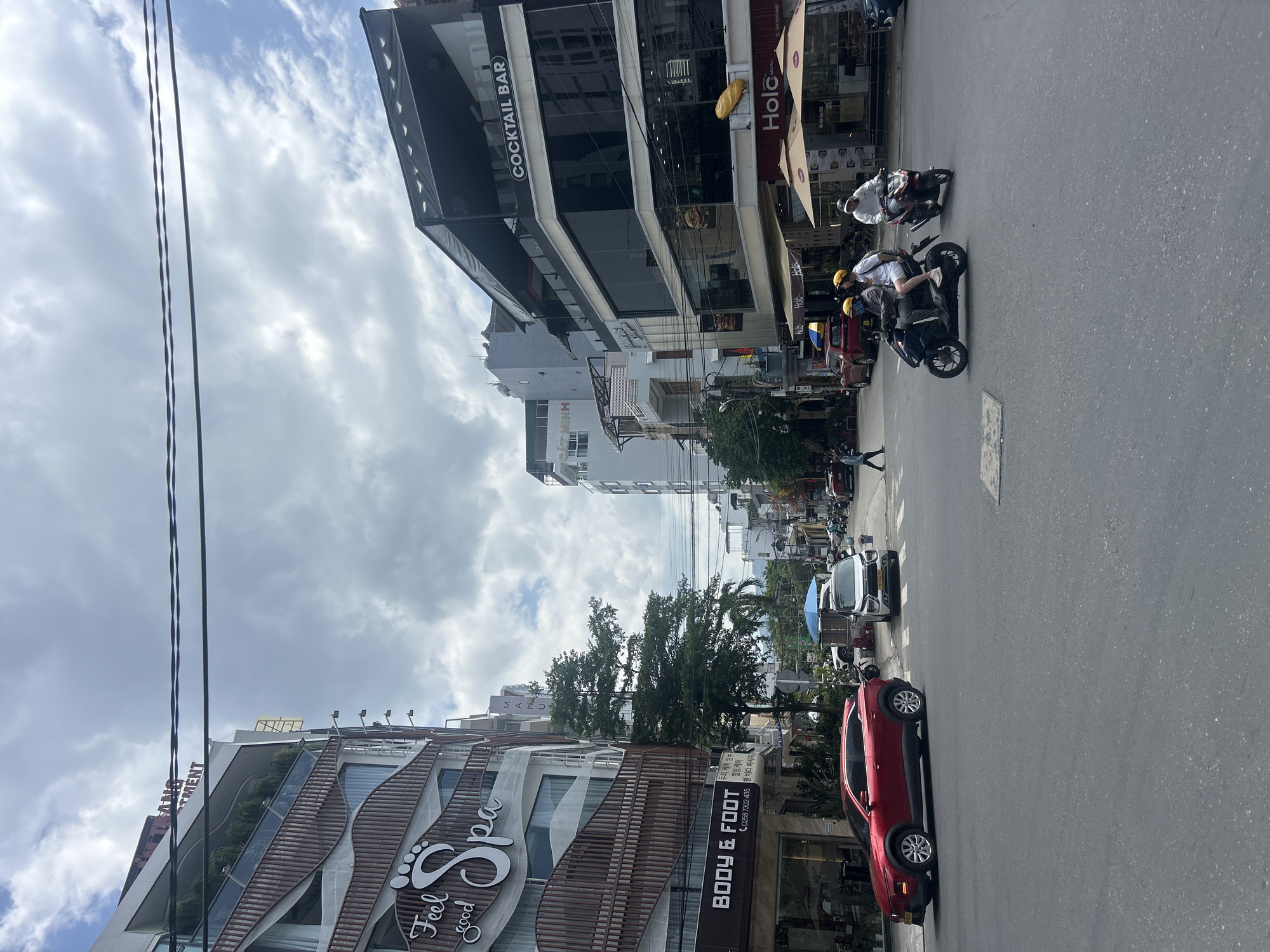 The image captures a bustling daytime street scene in Nha Trang Ward, Vietnam, characterized by multi-story urban buildings, various vehicles, and pedestrians under a partly cloudy sky.
On the left, a modern building with a distinctive wavy, wooden-slatted facade features a large sign reading "Feel good Spa" with a prominent footprint logo. Below this, a "BODY & FOOT" sign includes Korean text advertising "두피 케어 삼부" (Scalp care, Shampoo), "힐링케어" (Healing care), and "발 바디 마사지" (Foot body massage), alongside a phone number "0258 7302 435." Overhead power lines are visible against the sky.
On the right, another commercial building with dark glass windows displays a "COCKTAIL BAR" sign and a red banner for "Holc." The street itself shows active traffic and parking. Several motorbikes are present, including one with two helmet-wearing individuals and another with a single rider. A red sedan is parked closer to the foreground, and a white car is further down the street. A person can be seen walking on the sidewalk. Other partial text elements like "MINH," "MENT," and "MAU" are discernible on different buildings. The overall scene depicts a typical, active urban environment.
