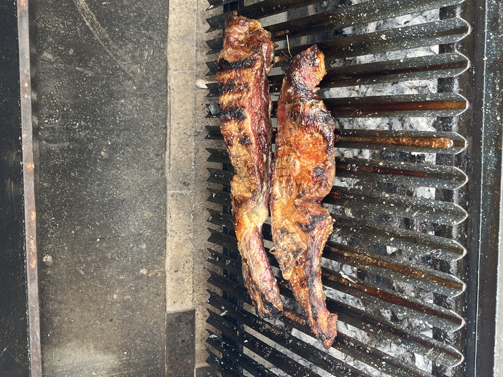 Two strips of grilled meat are centered on a grill. The grill is located near a concrete surface, possibly part of a parilla, a style of barbecue common in Buenos Aires, Argentina. The meat is visibly cooked, with grill marks. The grill is above embers of ash.