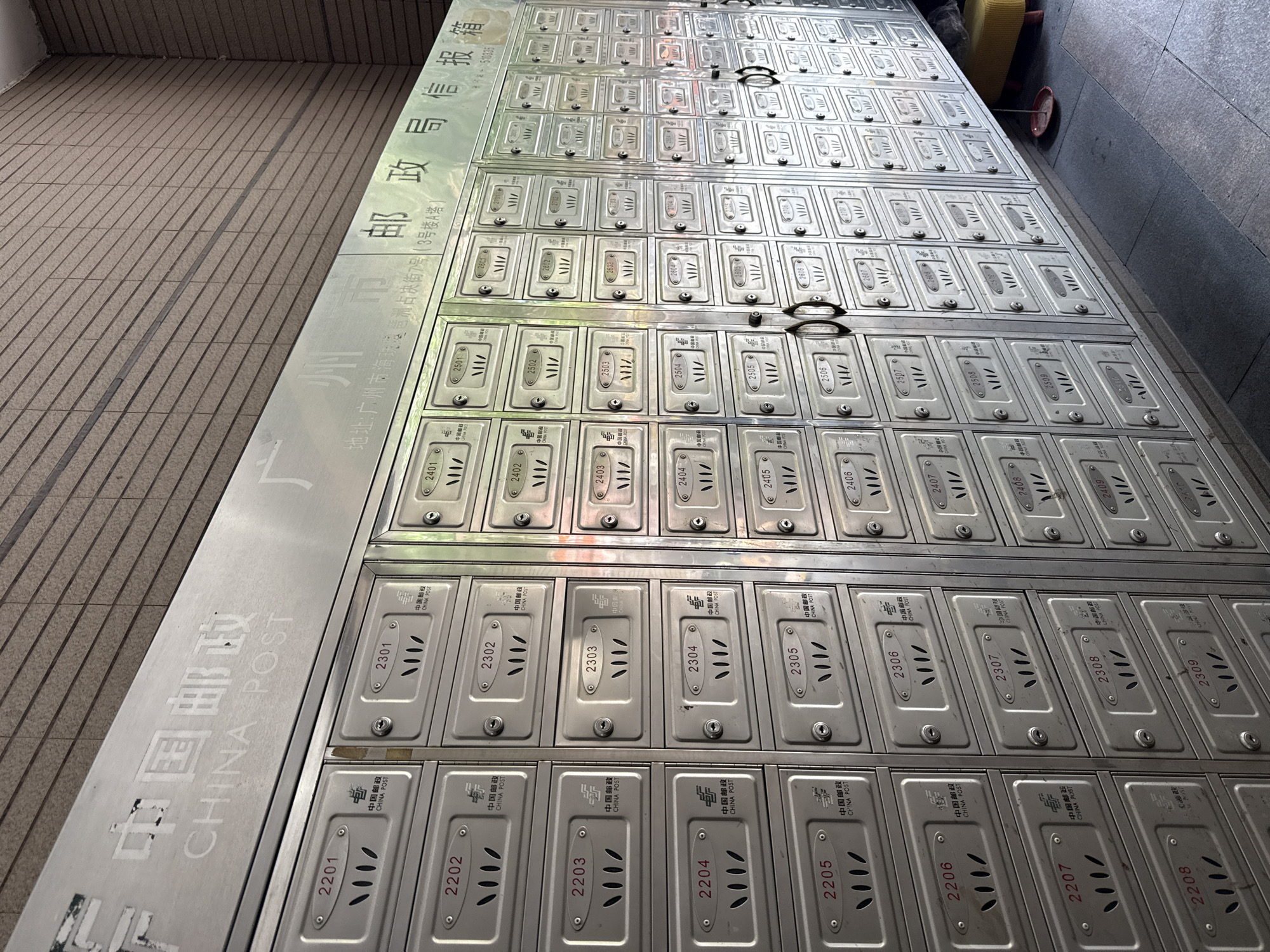 The image shows a large collection of metal mailboxes at the China Post office located at 7 Haizhu District, Guangzhou, China (Building A). The boxes are arranged in rows and columns and numbered starting with the 2200s up to 2600s. Each mailbox has a slot for mail and a locking mechanism. The sign above the mailboxes reads "China Post" in both Chinese and English.
