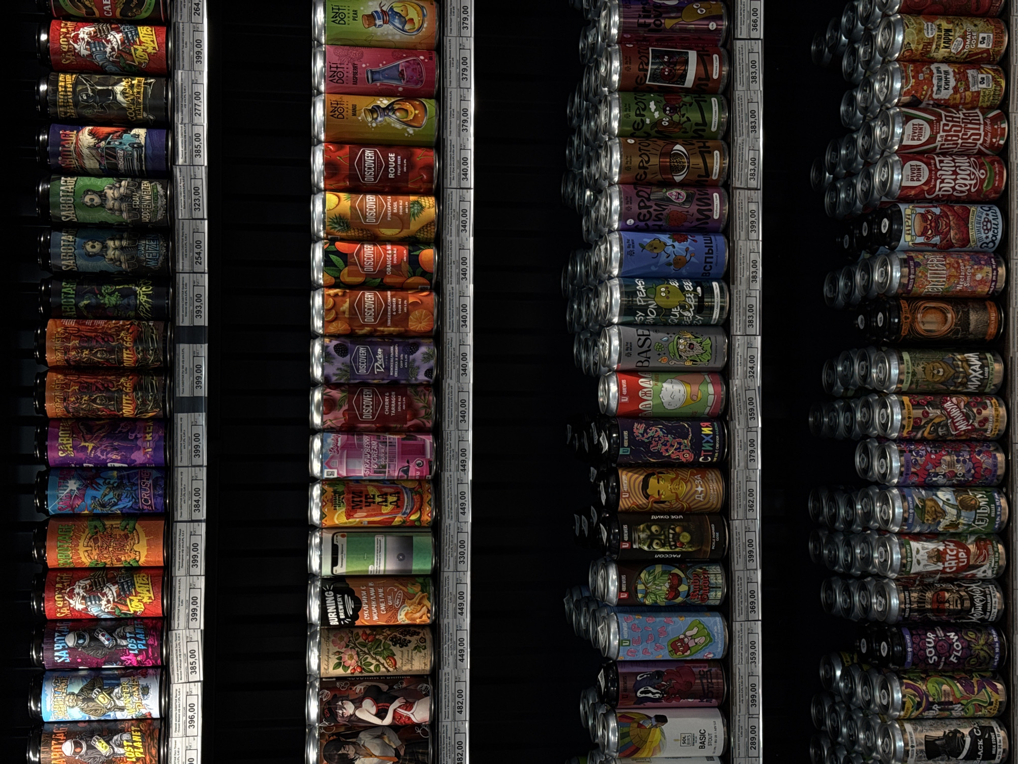 The image shows three shelves of canned beverages, presumably beer or soda, in what appears to be a retail setting. The shelves are densely packed with cans, each having a different label design and branding. Each can has a price tag adjacent to it. The first shelf from the left features cans with dark, illustrative labels in gothic and surreal styles, alongside beers from a brand called "Sabotage". The center shelf contains products from brands named "Discovery", "Anti Dot", and "Burning Brewery". The shelf on the right presents cans with more vibrant and diverse graphics and label names.