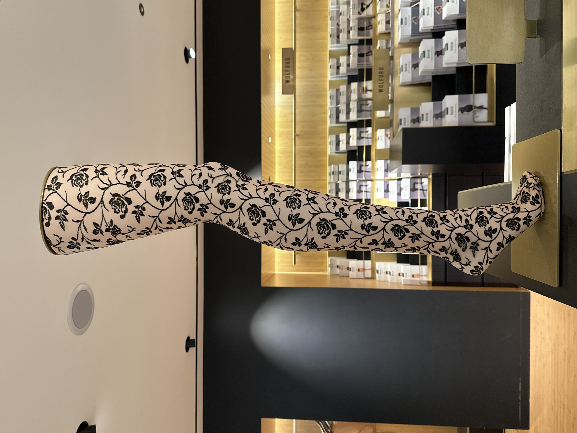 A single, disembodied leg wearing patterned stockings is displayed horizontally in a Wolford shop. The stocking is beige with a black floral pattern. The leg form is positioned within the store's interior, with a wall to the left and display shelves filled with Wolford products to the right. The store features a modern design with wood, black panels, and gold accents.