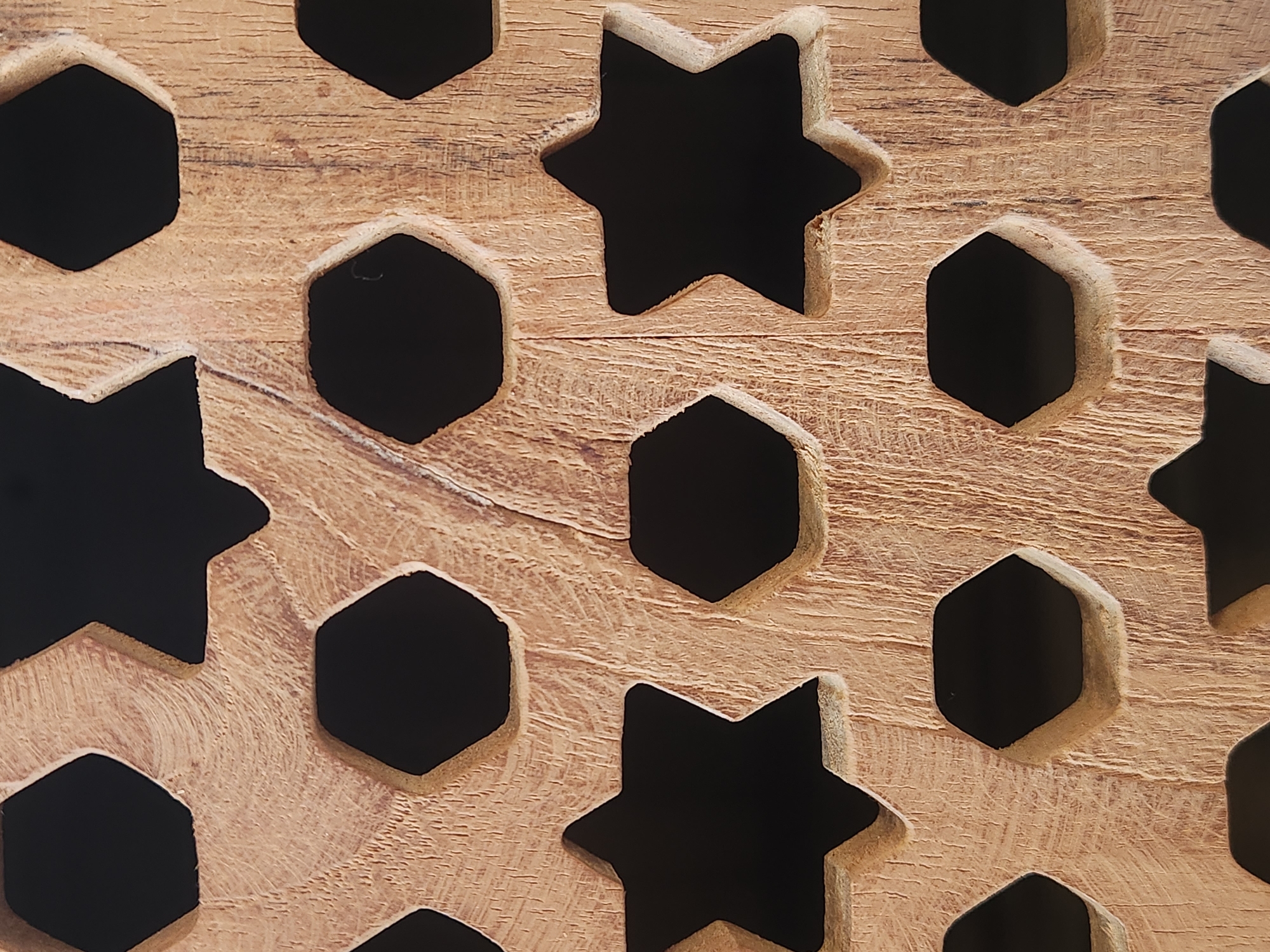 The image is a close-up shot of a wooden lattice with alternating hexagonal and star-shaped cutouts. The wood has a visible grain pattern with some imperfections and rough texture. The cutouts reveal a dark background, suggesting the lattice is either a screen or part of a larger structure. There are no people or animals present in the image. The overall impression is of a decorative element, possibly an architectural detail or a piece of furniture. There is no information available about the specific location of this object within the city of Maitland, United States of America.