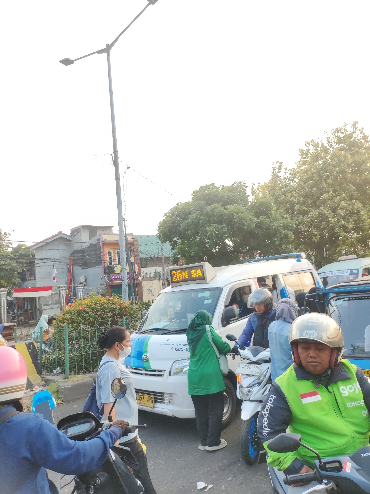 The image depicts a street scene in East Jakarta, Indonesia, characterized by traffic and pedestrian activity. A white van with blue and green accents, displaying the text "26N SA," is a central element, seemingly involved in public transportation. Several individuals are visible around the van, some on motorcycles and others on foot. One person, wearing a green jacket with "Gojek" branding and a Indonesian flag emblem, is seated on a motorcycle. Another individual in a blue jacket is also on a motorcycle, and a woman wearing a white mask and a light blue t-shirt is standing nearby. Green vegetation and trees form the background, with buildings and a street lamp visible in the distance. The lighting suggests it is daytime, possibly late afternoon due to the angle of the sun.