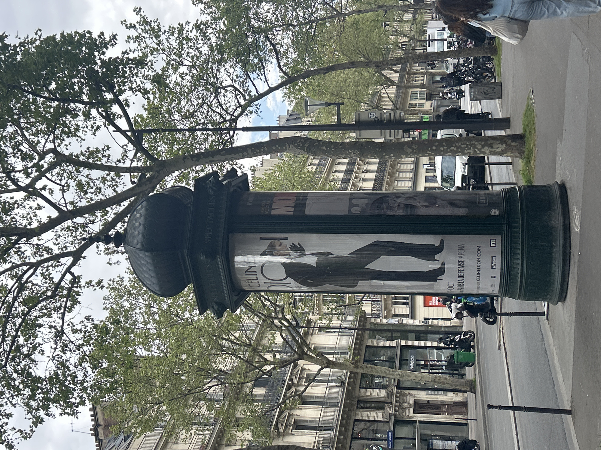 The image shows a street in Paris, France. In the foreground is a Morris column with a poster advertising a Celine Dion concert at the Paris La Défense Arena in October. The column is green with a black dome and ornate details. Behind the column are buildings with typical Parisian architecture, including balconies and wrought iron details. Trees line the street on both sides, with green leaves. Several motorcycles and scooters are parked along the side of the road. In the background, there are more buildings, trees, and a few people walking. The sky is overcast. The general environment seems to be a mix of residential and commercial.