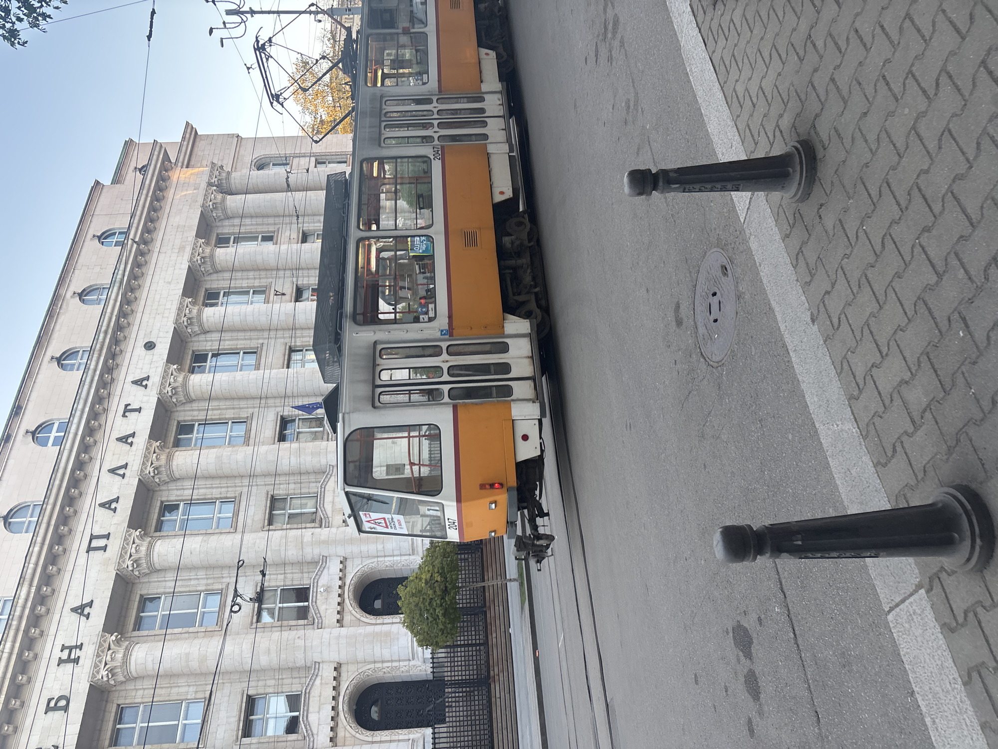 The image captures an urban street scene in Sofia, Bulgaria, featuring a tram running parallel to a large, imposing building under clear daylight. The tram, an older model with white and orange/yellow sections, displays the number "2047" prominently on its side. Its overhead power lines are visible against the bright sky.
To the left of the tram, a grand, multi-story building stands with a light-colored facade characterized by classical architectural details. It features numerous columns with ornate capitals, and a mix of rectangular and arched windows. Vertically inscribed in large Cyrillic letters on the building's side is the text "БЪЛГАPATA ПАЛАТА," which translates to "Bulgarian Palace," indicating a significant structure.
The street comprises a grey asphalt road with visible tram tracks, a white dividing line, and a patterned grey paved sidewalk. Two black, decorative bollards are positioned on the sidewalk, and a circular manhole cover is embedded in the asphalt. A small, leafy tree is visible behind a black metal fence in front of the building. The scene depicts a typical busy city environment, devoid of specific human activity or people.