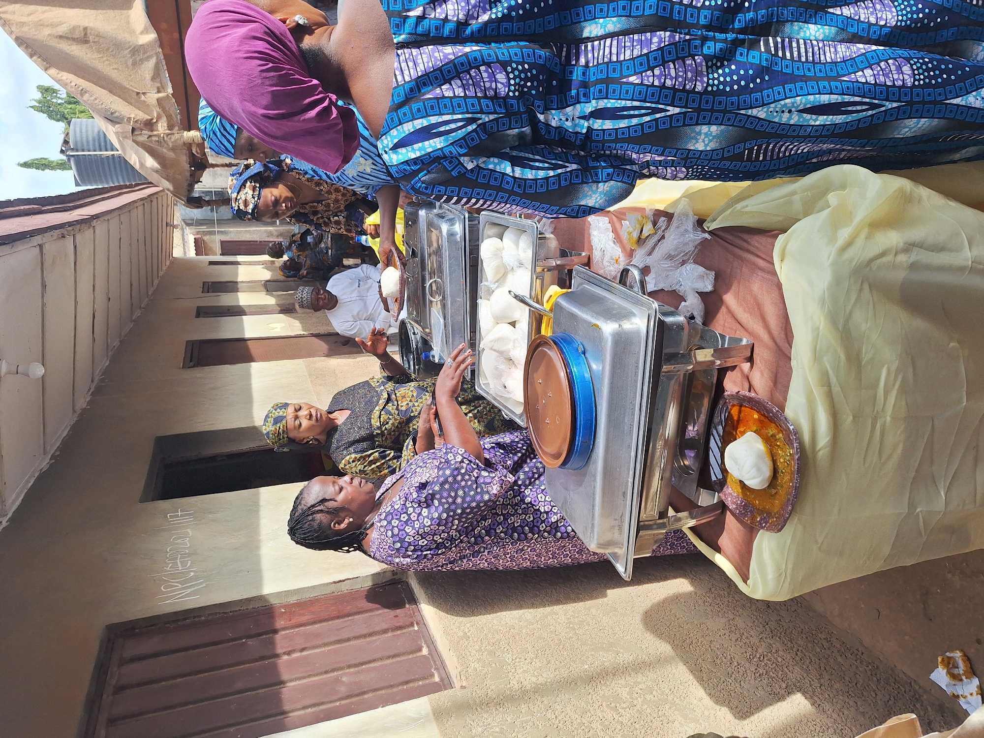 People stand in a line behind chafing dishes that contain food. The diners are wearing traditional Nigerian dress and some are wearing head coverings. A man in white stands in the line, speaking with his hands. A building with doors and windows is in the background. The writing "NPCEABSOLUT17" is on the wall. The location is Sabon Tasha, Nigeria.