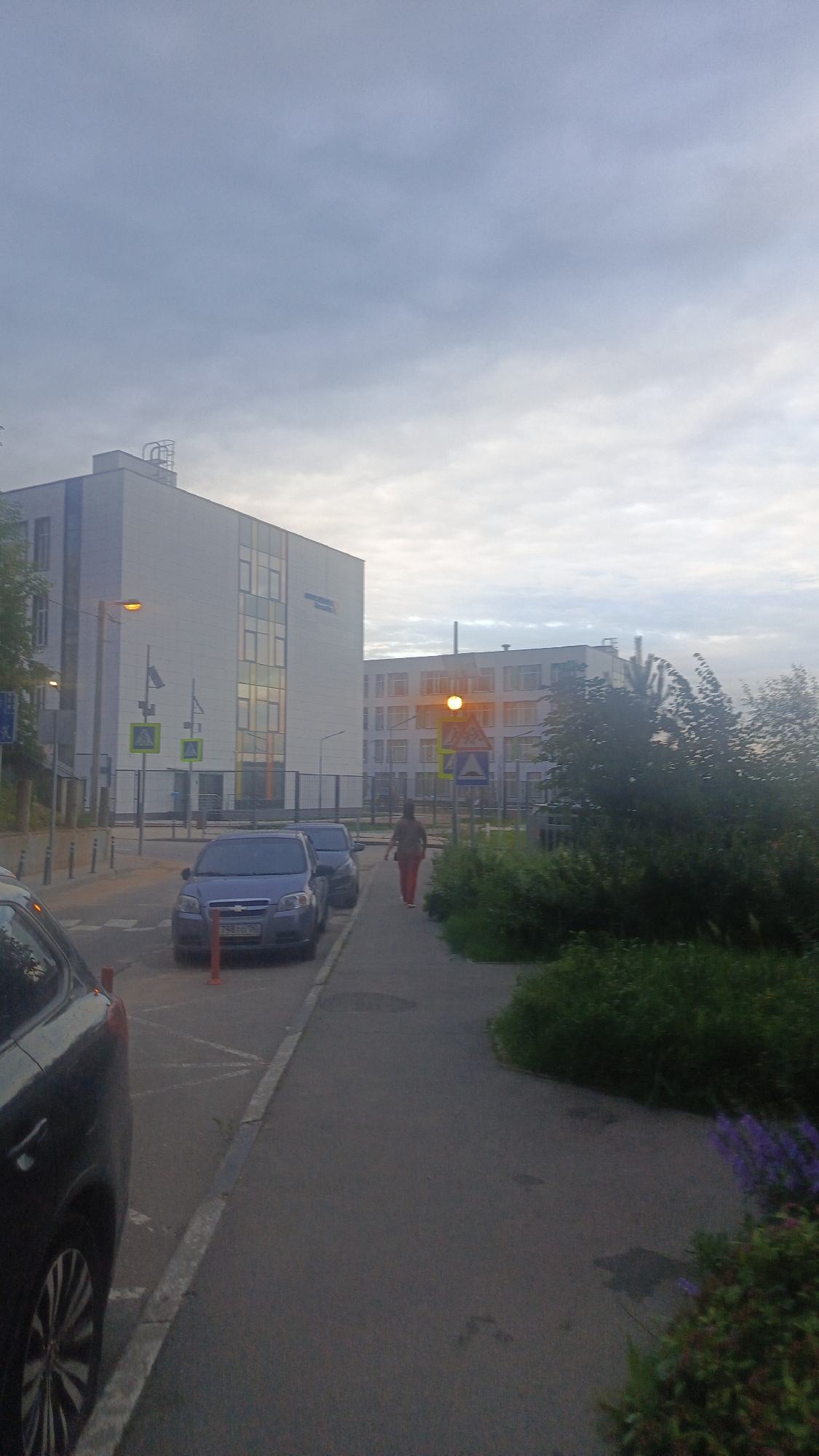 The image depicts a street scene in Zvenigorod, Russia. In the foreground, a dark-colored car is parked on the left side, with its rear visible. The street continues past parked cars, including a blue sedan. A pedestrian, wearing reddish-brown pants and a dark top, walks away from the camera along the sidewalk. Across the street, two large modern buildings are visible. The building on the left is white with multiple windows and some blue accents. The building on the right also appears to be a school or institutional building, with numerous windows and some architectural details. A traffic light showing amber is visible in front of the right building. Several street signs are present, including yellow diamond-shaped warning signs. The sky is overcast with clouds. The scene appears to be on a street with a sidewalk and a parking area.
