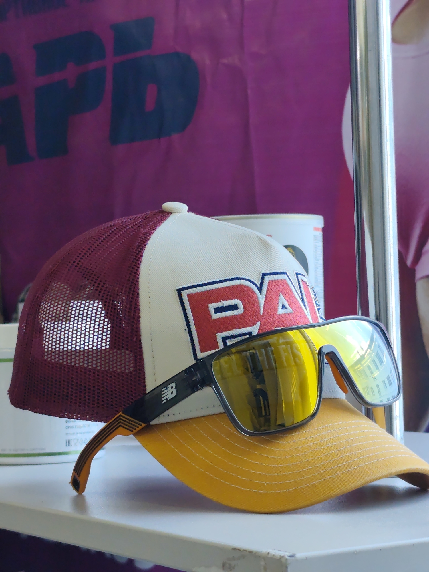 A close-up, eye-level shot features a baseball cap and sunglasses positioned on a white surface, likely a shelf or table, against a blurred background. The cap has a maroon mesh back and a cream-colored front with red lettering that reads "PA" outlined in blue. A yellow visor is attached to the cap. Resting on the cap are a pair of black-framed sunglasses with mirrored yellow lenses, reflecting the surrounding environment. The sunglasses have orange accents on the temples and a New Balance logo on the left temple. To the left of the cap, a white cylindrical object, possibly a container, is partially visible. The background is a deep magenta fabric with a large, dark blue graphic, seemingly an abbreviation or logo. A vertical silver pole stands to the right of the subjects. The scene appears to be an indoor setting with artificial lighting, based on the reflections in the sunglasses. The image is sharp in the foreground and progressively softer in the background. The location in Chaikovsky, Russia is not identifiable from the visual elements present in the image.