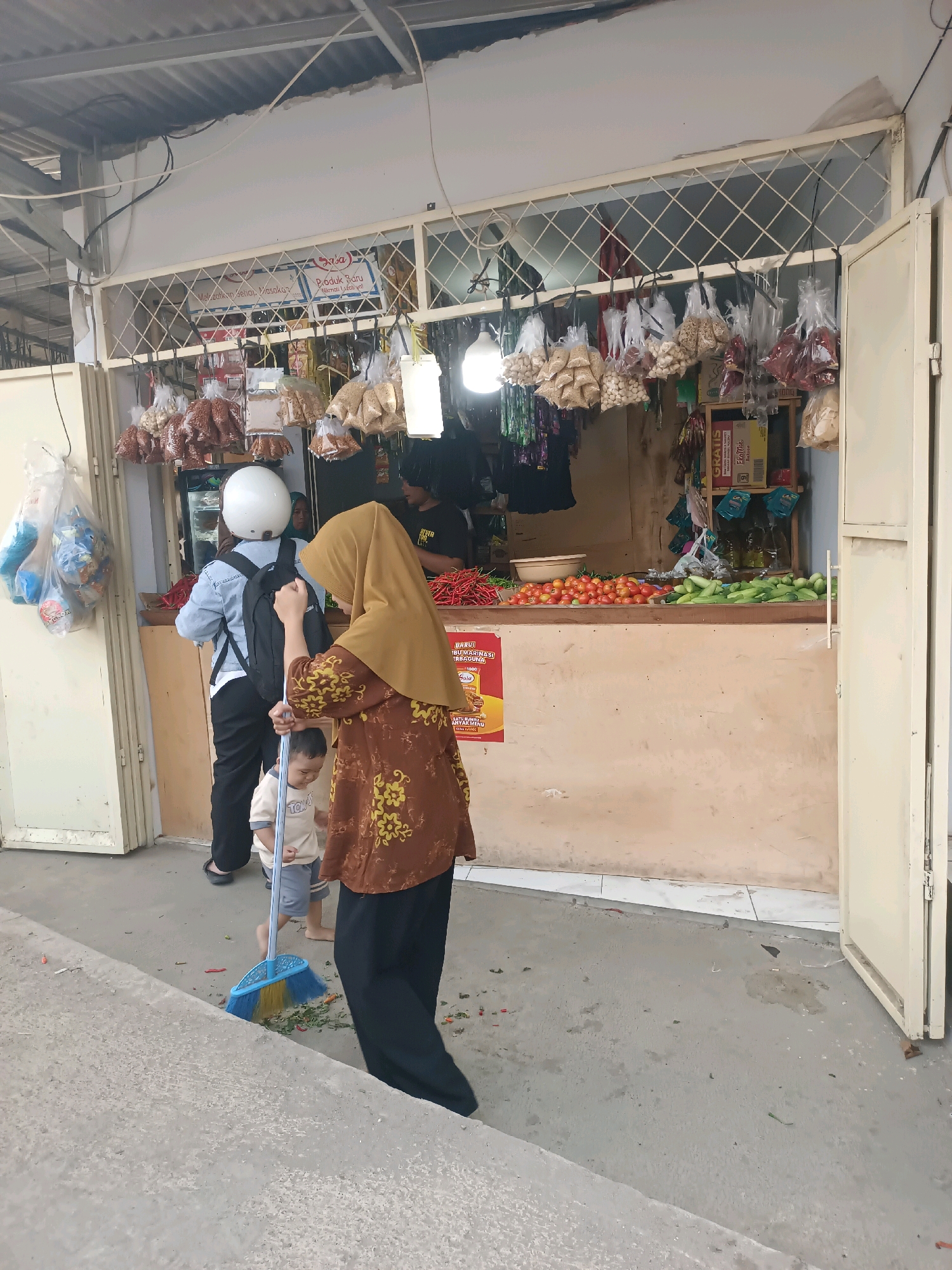 The image depicts an outdoor market stall in Karawang, Indonesia. In the foreground, a woman wearing a brown and yellow patterned tunic and a yellow hijab is sweeping the ground with a blue and yellow broom. A young child in a light-colored shirt and shorts stands nearby. Behind them, a person in a denim jacket and white helmet is facing away from the camera, with a black backpack. The market stall features shelves laden with various goods, including bags of spices or nuts hanging from the ceiling, and fresh produce like tomatoes and cucumbers displayed on the counter. A red chili pepper is visible on the counter. The stall has an open front with white doors and a metal grate. The overall environment appears to be a typical street market scene in Indonesia.
