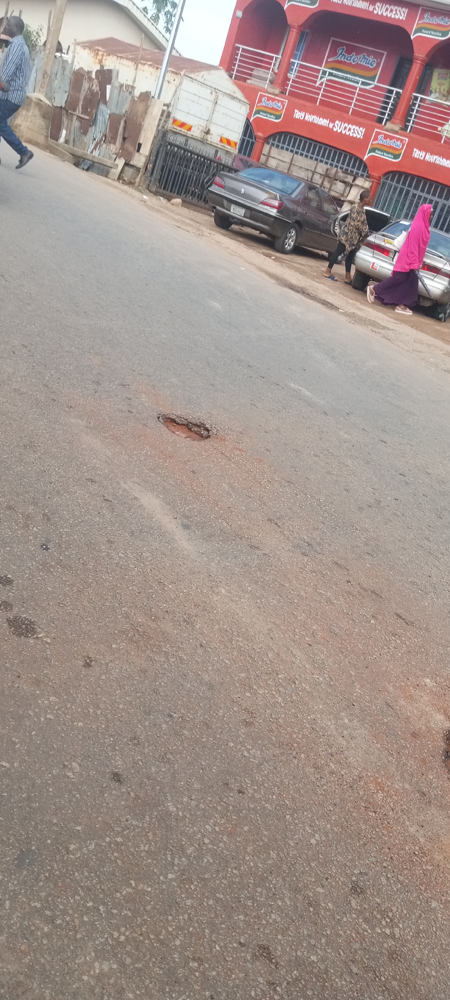 The image shows a street scene in Jos, Nigeria, with a significant pothole in the asphalt road surface. The pothole is roughly oval-shaped and reveals exposed sub-layers of the road. Several individuals are present: one man is walking on the left side of the road. There are two other persons visible near parked cars, one wearing a floral dress and the other wearing a pink headscarf. In the background, there are at least two parked cars with visible license plates. Behind the parked cars is a red multi-story building advertising "Indomie," a brand of instant noodles, with the slogan "Tasty Nourishment or SUCCESS!". Other objects present include a white truck and a metallic structure to the left of the truck.
