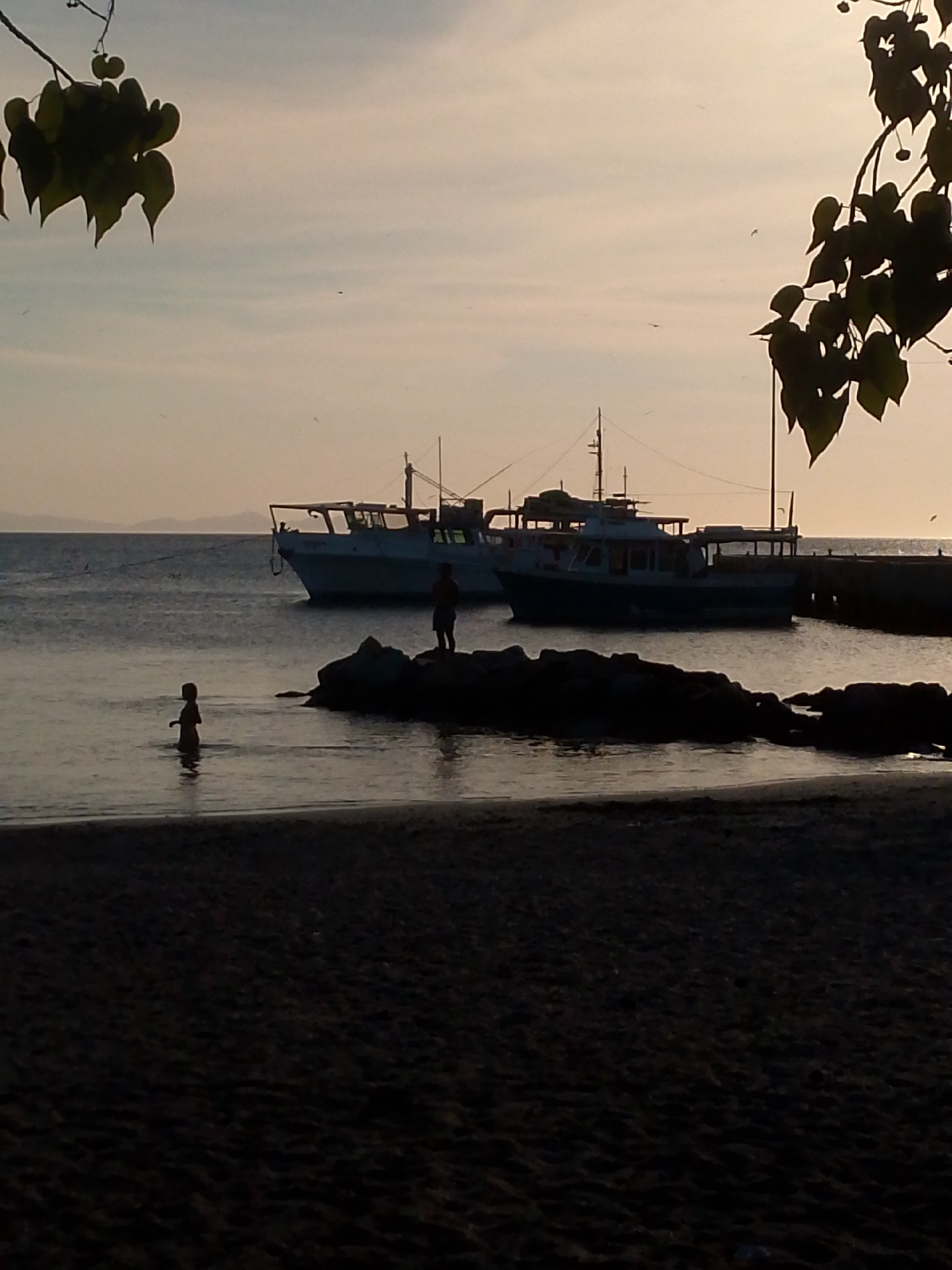 The image shows a beach scene at dusk in Juan Griego, Venezuela. In the foreground is a dark sandy beach. In the middle ground, the sea reflects the fading light. Two silhouettes of people are visible: a child wading in the water near the beach and an adult standing on a rock formation further out. Two boats are docked near a pier in the background. Leaves of trees are visible in the top corners of the frame. In the far background, a hazy land mass is visible on the horizon.