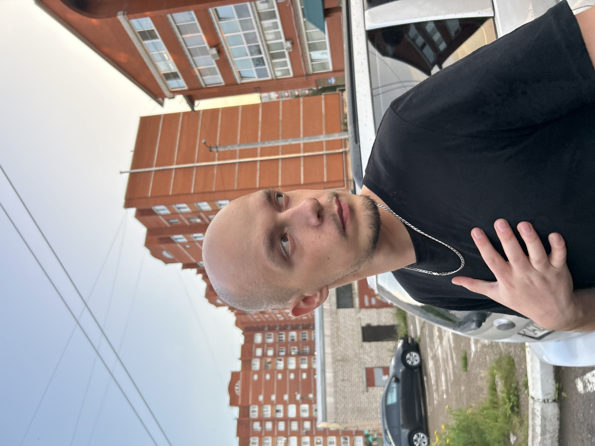 A young bald man with a goatee and a silver chain necklace stands in a parking lot. He is wearing a black t-shirt and has his left hand placed on his chest. Behind him, several multi-story apartment buildings constructed of orange brick are visible. A street light stands in front of one of the buildings, and a dark-colored sedan is parked in the background. The scene appears to be set in the city of Chaikovsky, Russia.