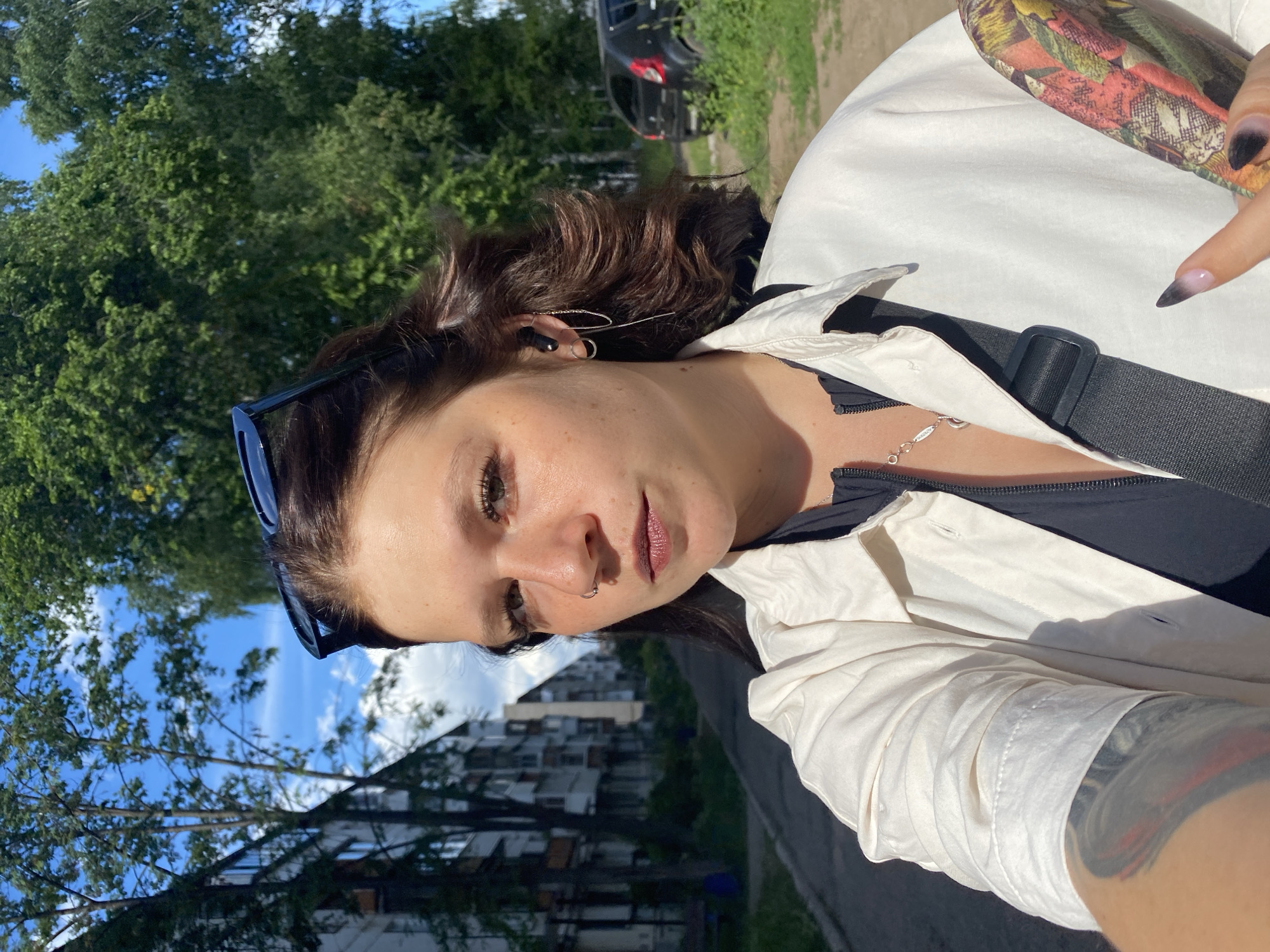 In Chaikovsky, Russia, a young woman is captured in a self-portrait outdoors. She is facing the camera with sunglasses perched on her head and her dark hair windswept. She wears a white zip-up jacket over a black top, a silver necklace, and hoop earrings with chains. A nose ring is visible, and her left arm displays a colorful tattoo. She holds a patterned object in her right hand, and her nails are painted in a gradient of black and white. In the background, apartment buildings line a paved walkway, with trees and a dark car on the right. The sky is a bright blue with scattered clouds.