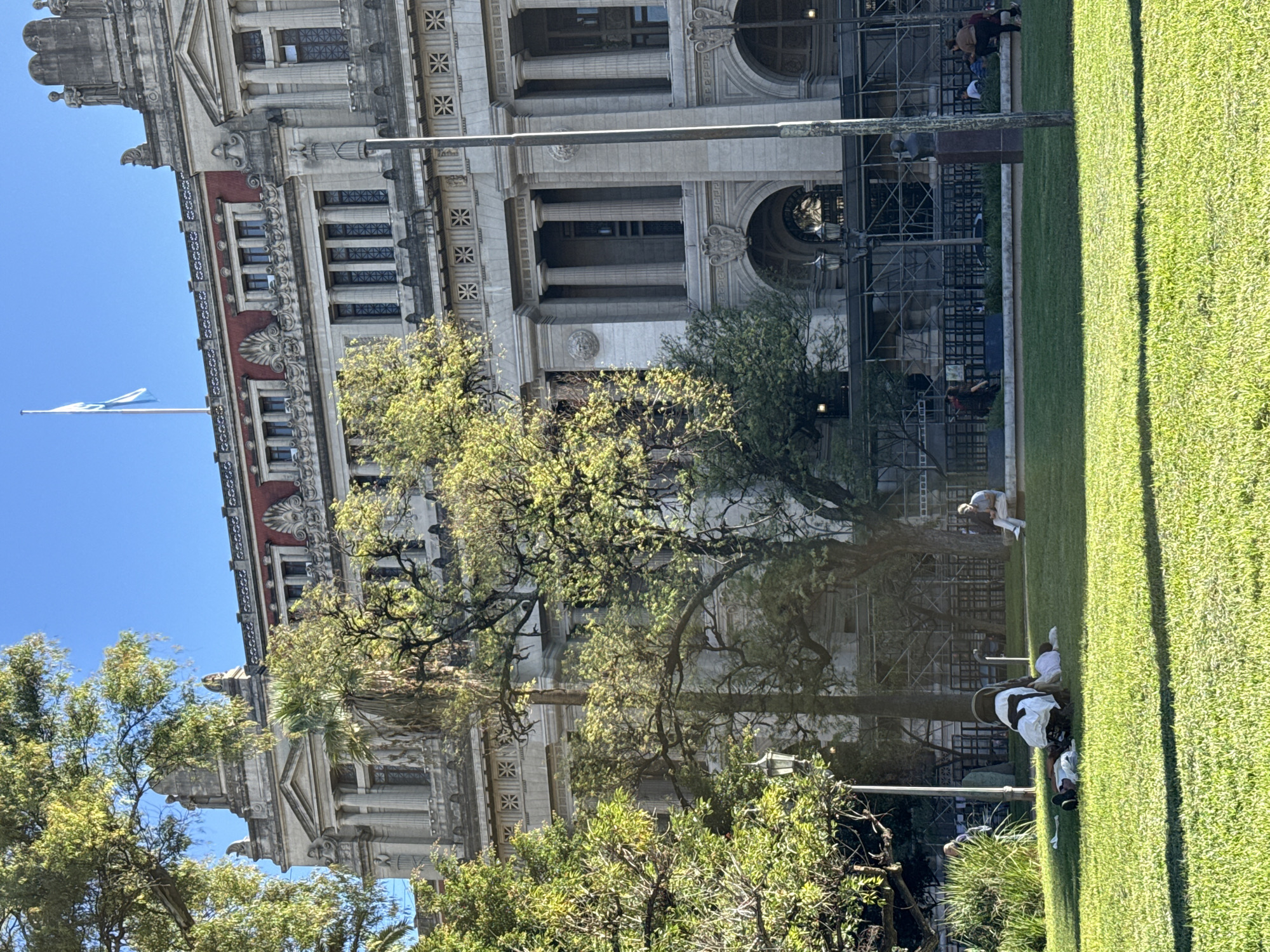 This image depicts a sunny day in what appears to be a public square or park in Buenos Aires, Argentina, given the context.
In the background stands a large, ornate building exhibiting classical architecture. It features multiple stories, grand columns, decorative cornices, intricate carvings, and arched windows. The building is primarily constructed from light-colored stone, with sections of red brickwork adding contrast. Scaffolding is visible on parts of the building, particularly around the arched lower sections, suggesting ongoing renovation or maintenance work.
To the left, against a clear blue sky, a light blue and white flag, characteristic of Argentina, flies from a tall pole.
The mid-ground and foreground are dominated by a lush, well-maintained lawn of green grass. Several trees with bright, verdant foliage are scattered throughout, some partially obscuring the building's facade.
Several people are present in the scene, mostly relaxing on the grass. Some individuals are seen lying down, others are sitting, and a few are standing or walking near the edge of the grassy area or close to the building's scaffolding. Their casual postures indicate a leisurely activity.
The time of day appears to be late morning or early afternoon, characterized by bright sunlight casting clear shadows, indicating a sunny and pleasant weather condition. No discernible text is visible on the building or within the scene. The overall atmosphere is tranquil and relaxed.