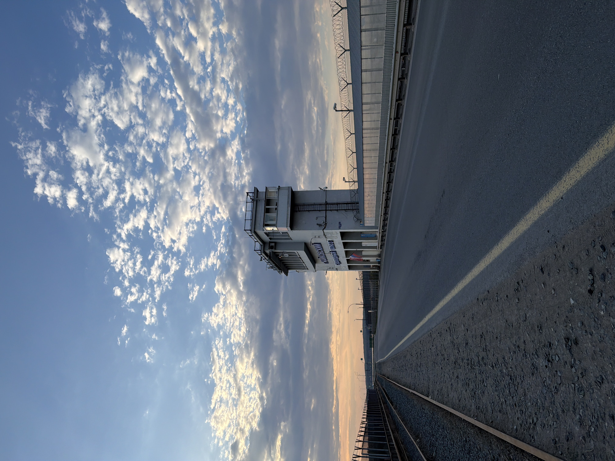 A daytime, rotated shot shows a building with the name "Чайковский Завод" (Chaikovsky Plant) in Chaikovsky, Russia. The sky features a variety of clouds. There is a fence with barbed wire along the top. A road is visible next to the building, as well as railroad tracks on the other side of the road. Street lights are in the background.