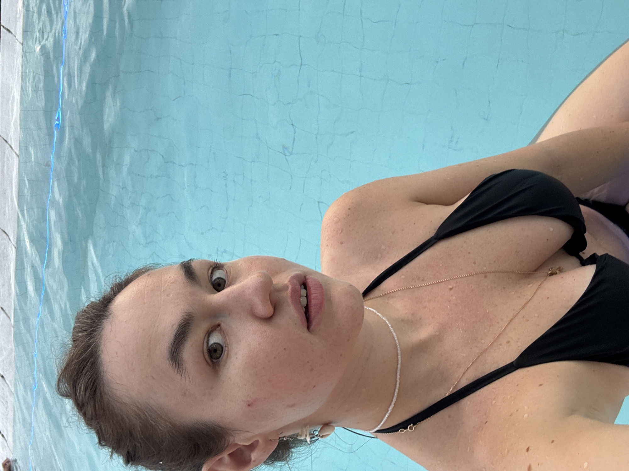 A young woman is captured from the chest up, posing for a selfie in a swimming pool. She is wearing a black bikini with thin straps. Her hair is pulled back, revealing her face and shoulders. She has light skin with freckles and is looking directly at the camera with a neutral expression. She is wearing a seashell earring on her left ear and two necklaces. The background is a bright blue swimming pool with visible tile lines. The water appears to be clear and shallow. The image is well-lit and focused on the woman. The location is identified as Karon, Thailand.