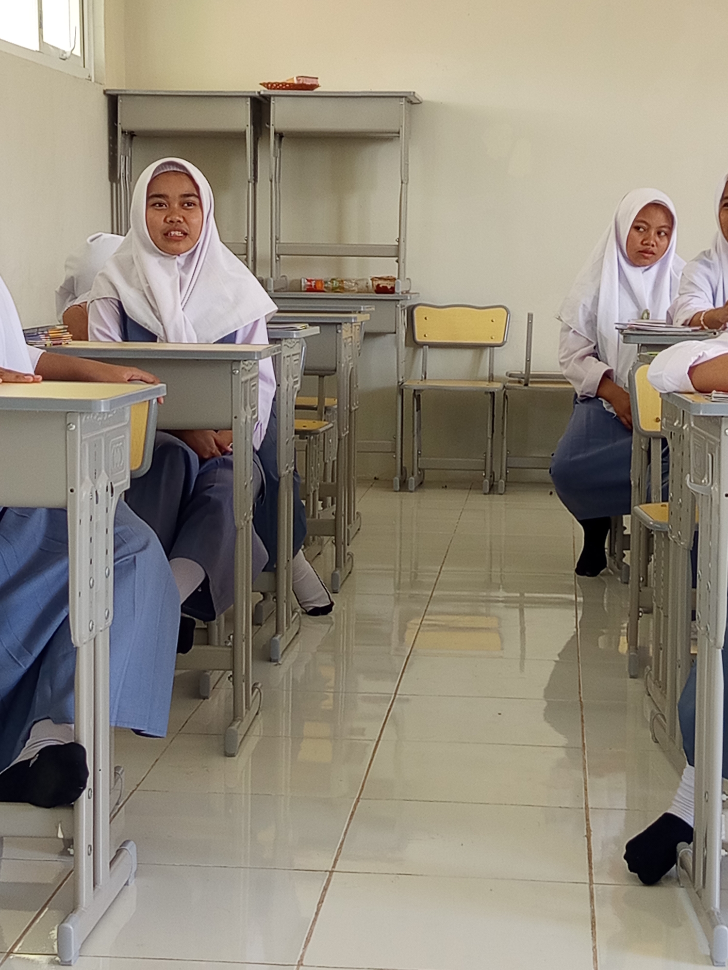 In a classroom in Lumutan, Indonesia, several young women wearing white hijabs and blue school uniforms sit at desks. The room has a tiled floor and bright lighting. One student in the foreground on the left is looking forward with her mouth slightly open. Other students are visible in the background, some looking away from the camera. Desks and chairs are arranged in rows. Shelving units are against the back wall, holding various items. The environment suggests a typical school setting during a lesson.