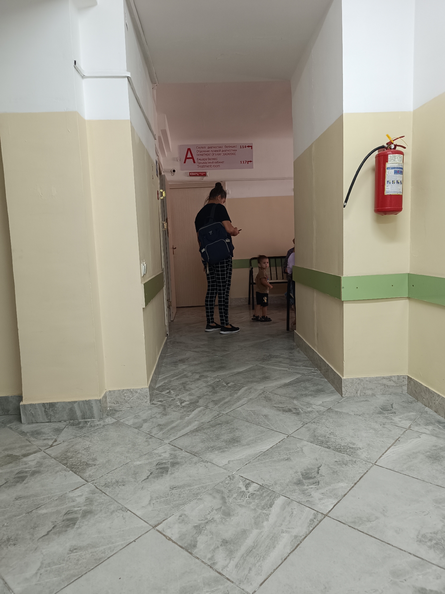 The image depicts an indoor hallway within a building, possibly a medical facility, given the signage. A woman with dark hair, wearing a black t-shirt and checkered pants, is standing with her back to the camera, holding a phone and a dark bag over her shoulder. To her left, a young child in a brown t-shirt and shorts stands facing towards a doorway. Further in the background, another person is partially visible sitting on a bench. On the wall to the right, a red fire extinguisher is mounted. The hallway features cream-colored walls, a green handrail, and a tiled floor with a marble-like pattern. Above a doorway on the left, a sign with Cyrillic and English text, including "DEPARTMENT OF X-RAY DIAGNOSIS," is visible. Based on the provided context, this scene is located in the city of Taraz, Kazakhstan.