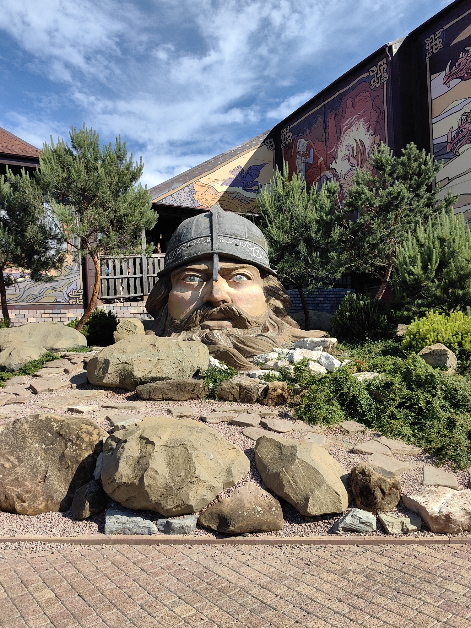 The image captures a large, realistic sculpture of a man's head with a helmet, emerging from the ground in what appears to be an outdoor exhibition or park area. The sculpture features a stern expression, a long beard, and light blue eyes. It is surrounded by large rocks, gravel, and low-lying green foliage. Behind the sculpture, there is a building with a large mural on its exterior wall, depicting fantastical scenes with figures and dragons. The architecture includes wooden elements and a tiled roof. The sky above is blue with scattered white clouds, suggesting it is a clear day. The foreground shows a brick-paved walkway. This scene is located at "Skver Nauki" (Science Park) in Sochi, Russia, a place known for its artistic installations and thematic areas.