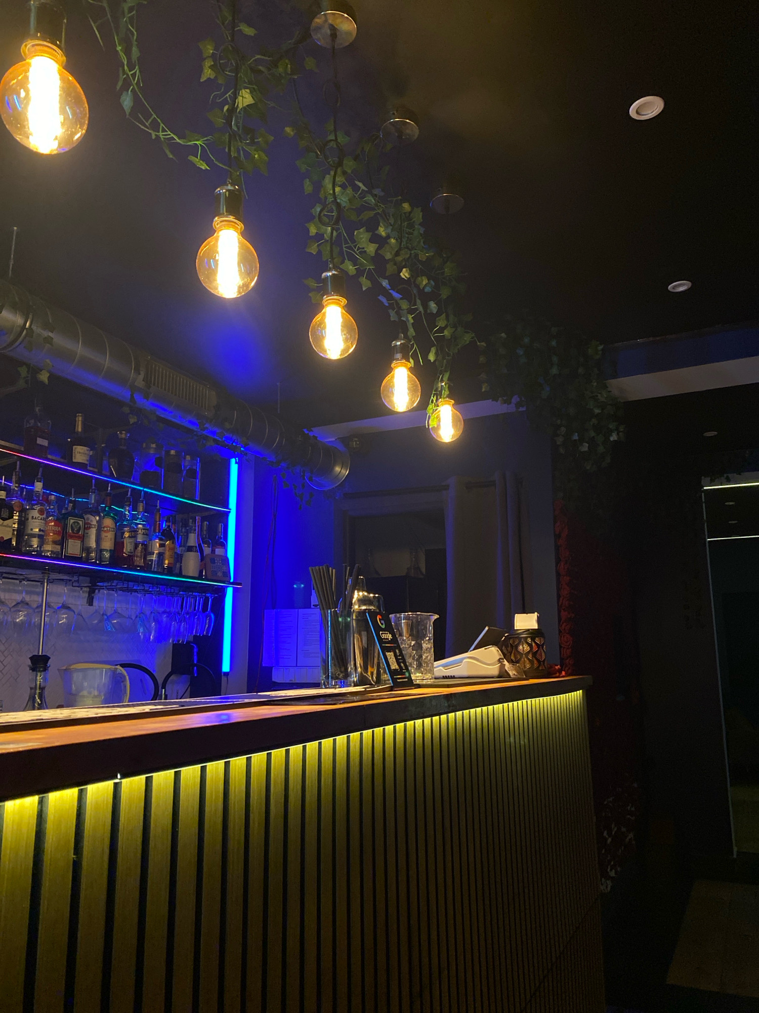The image depicts the interior of a bar, featuring a well-stocked bar shelf illuminated by blue and purple LED lights. Hanging pendant lights with exposed incandescent bulbs are suspended from the ceiling amidst artificial greenery. The bar counter is made of wood, with a vertical slatted design on its front, lit by a warm yellow underglow. Several bottles of liquor are visible on the shelves, along with glassware and bar tools on the counter. The overall ambiance is dark and atmospheric, suggesting a nightlife setting. This image is of a bar located at 74 Stanisława Dubois Street, Gliwice, Poland. There are no people or discernible actions taking place in the scene.