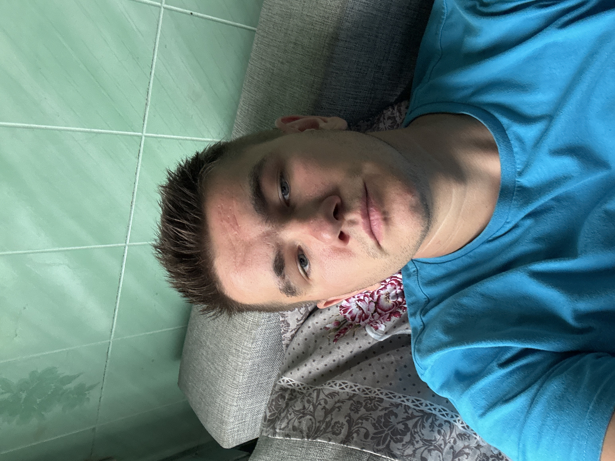 A young Caucasian male is shown from the chest up, resting against a grey couch with a patterned textile. The male has short, spiked brown hair, blue eyes, and is wearing a bright blue t-shirt. He has light stubble on his face and appears to be looking directly at the camera. Behind him, a wall is visible, tiled in a light green color. The lighting suggests an indoor environment, possibly a home. No specific location in the city of Krasnoe, Russia, can be identified from the image.