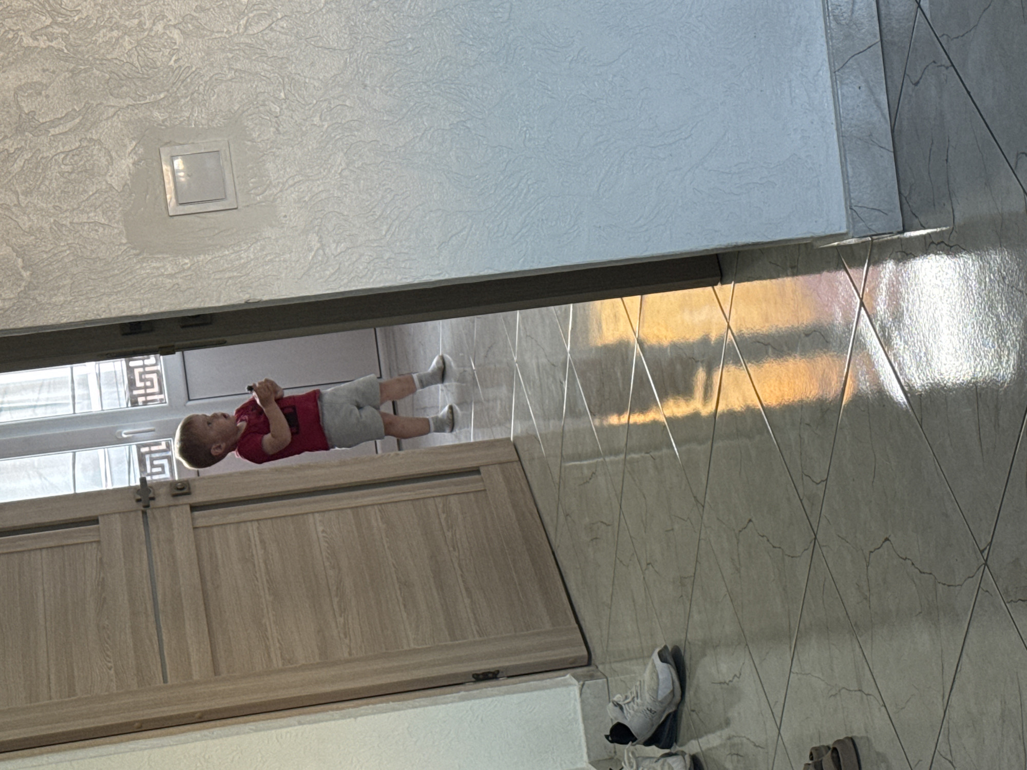 A young child wearing a red t-shirt and grey shorts stands in a hallway. The child is holding a dark object in their right hand. To the left, a wooden door is ajar, revealing part of a window with decorative bars. A pair of white athletic shoes with black accents rests on the tiled floor in the foreground. The hallway floor is tiled with a diamond pattern and reflects the sunlight streaming in from the window, creating a warm, orange glow. To the right, a textured wall features a white light switch. The scene appears to be indoors in a residential setting.