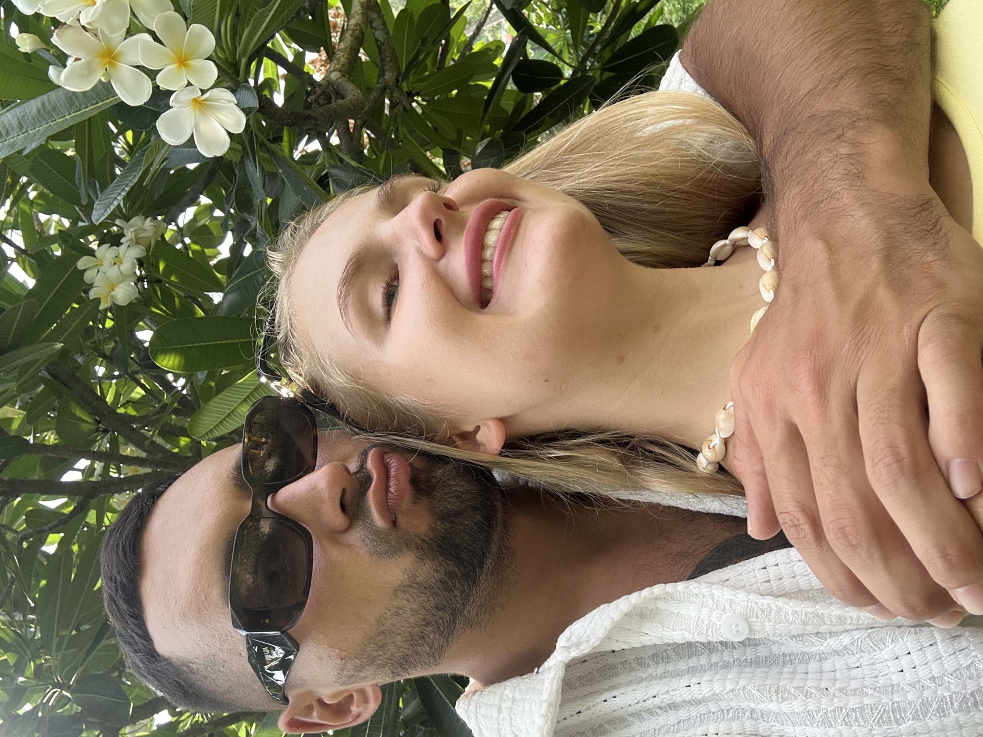 The image shows a close-up, low-angle shot of a couple embracing outdoors, surrounded by lush greenery and white frangipani flowers. The man is in the foreground, wearing sunglasses and a white textured shirt. He has a beard and is looking up. The woman is behind him, leaning her head on his shoulder, with her eyes closed and a smile on her face. She has blonde hair and is wearing a shell necklace. The scene appears to be set in a tropical or sub-tropical location, possibly Vietnam, given the frangipani flowers and dense foliage. The lighting suggests it is daytime, with sunlight filtering through the leaves. The couple appears relaxed and happy, enjoying an intimate moment together. No visible text is present in the image.