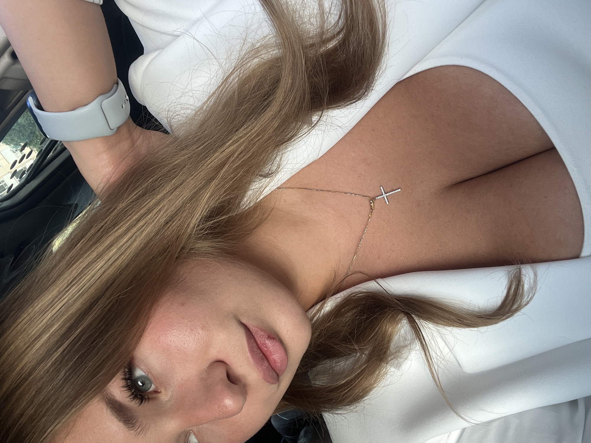 A woman with light brown hair and fair skin is visible from the chest up. She is wearing a white jacket, and a delicate gold necklace with a diamond cross pendant rests against her skin. A gray Apple Watch is on her left wrist. The background suggests she is in a car, with glimpses of the outside environment, possibly trees, seen through the window. The location is unknown.