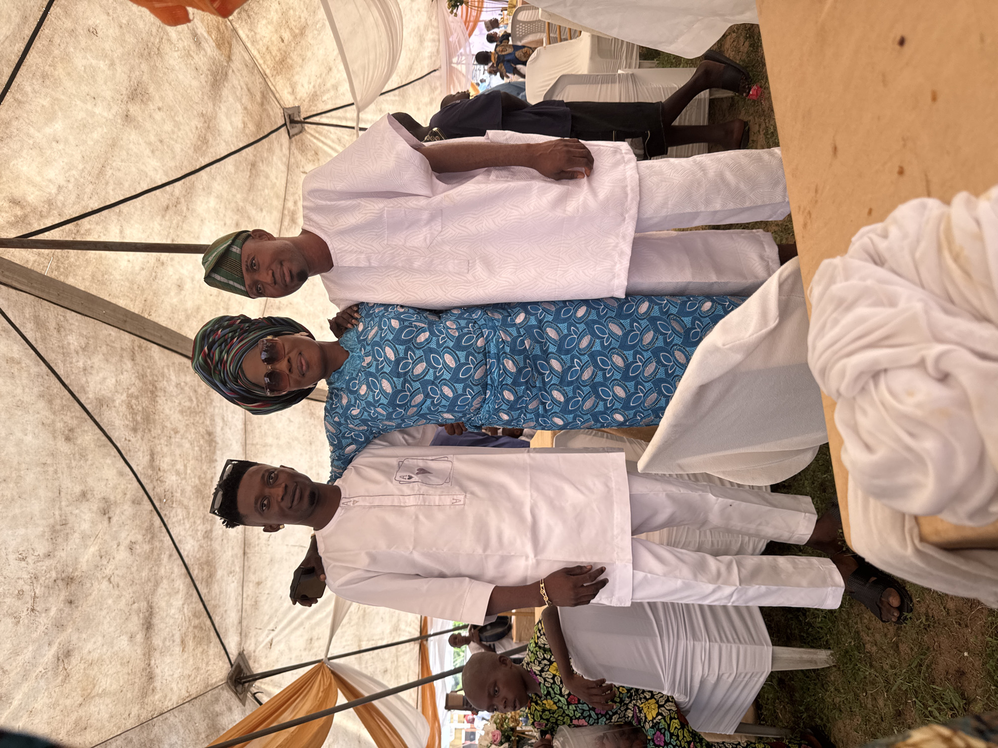 The image shows three people posing for a photo under a tent. Two men, dressed in white traditional attire, stand on either side of a woman wearing a blue patterned dress and a headscarf. The man on the left has sunglasses pushed up on his head and a gold bracelet on his wrist. The man on the right wears a green and white checkered cap. A young child in a colorful floral outfit is seated to the left of the frame, looking away from the camera. The environment appears to be outdoors, with a tent providing shade, suggesting an event or gathering, possibly in the city of Badagry, Nigeria, given the context. Chairs and tables are visible in the background, along with other people and some decorations. The overall scene conveys a sense of celebration or a social occasion.
