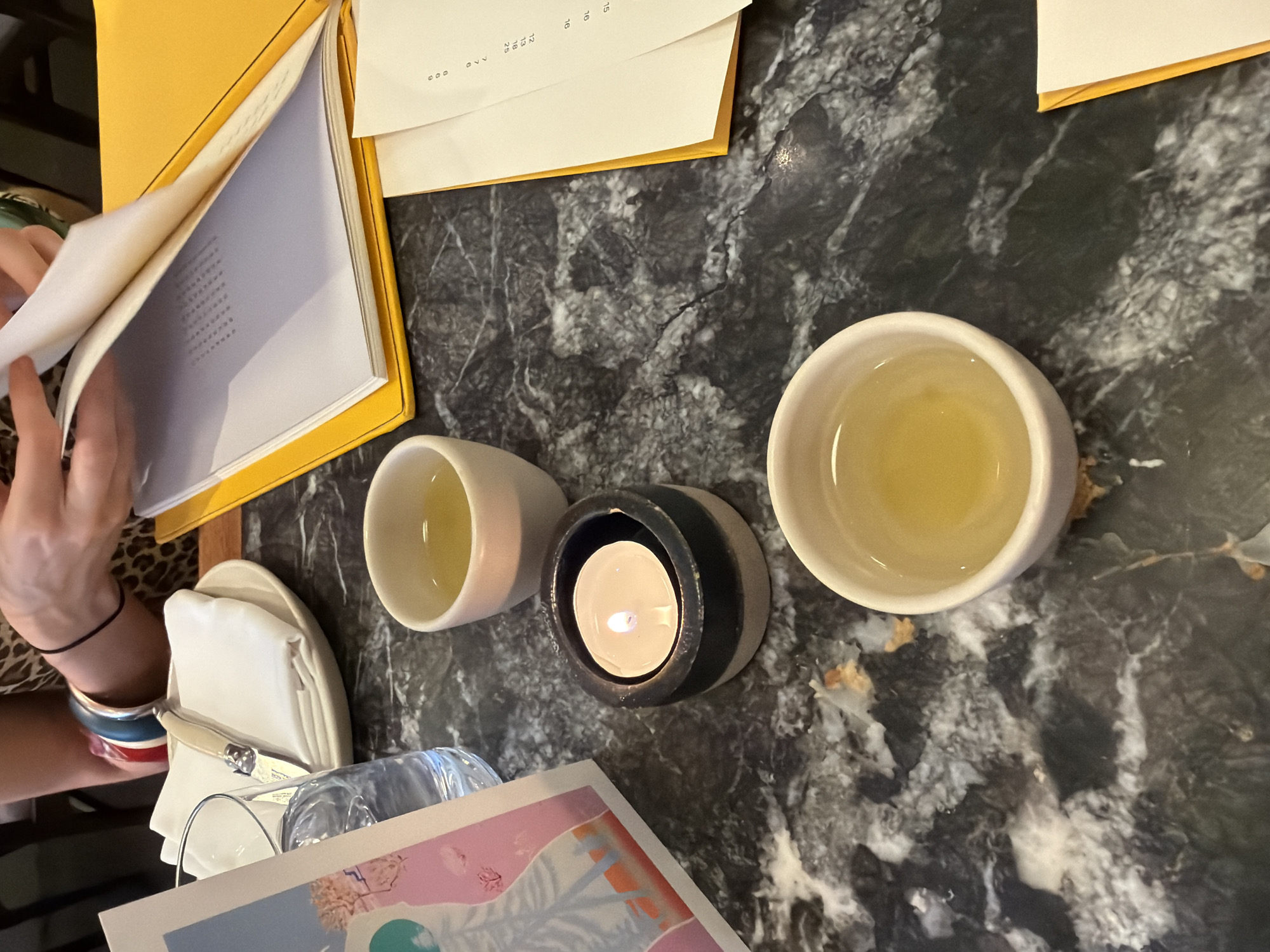 A close-up, eye-level shot captures a dimly lit restaurant table with a dark, marbled surface. Three small, white teacups containing a light amber liquid are arranged in a triangular formation. A small, black and gray ceramic candle holder with a lit tea light sits between two of the cups. To the left, a partially visible glass of water is next to a small, white plate with a folded white napkin and a knife with a light-colored handle. A colorful postcard with an abstract illustration of a figure and pink floral elements is propped up on the left side of the frame. In the background, a person's arms are visible, wearing a leopard-print top and colorful bracelets, and they appear to be holding and looking at a book or menu with a bright yellow cover. The overall ambiance suggests an intimate dining experience in London, United Kingdom.