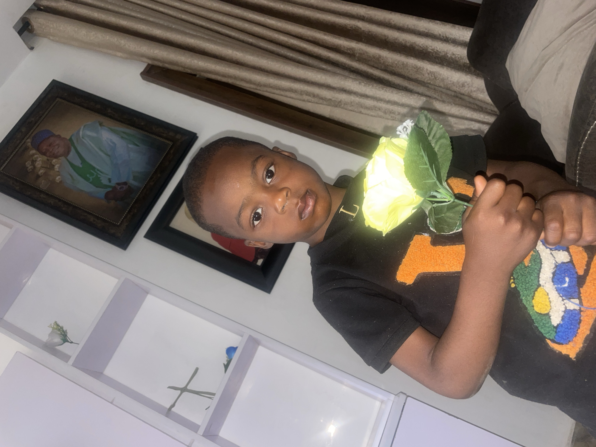 A young child holds a yellow rose. Behind the child are two framed portraits hanging on the wall and a white shelf with floral decoration. The location is likely indoors but there is no verifiable evidence to determine if it is in Jos, Nigeria.