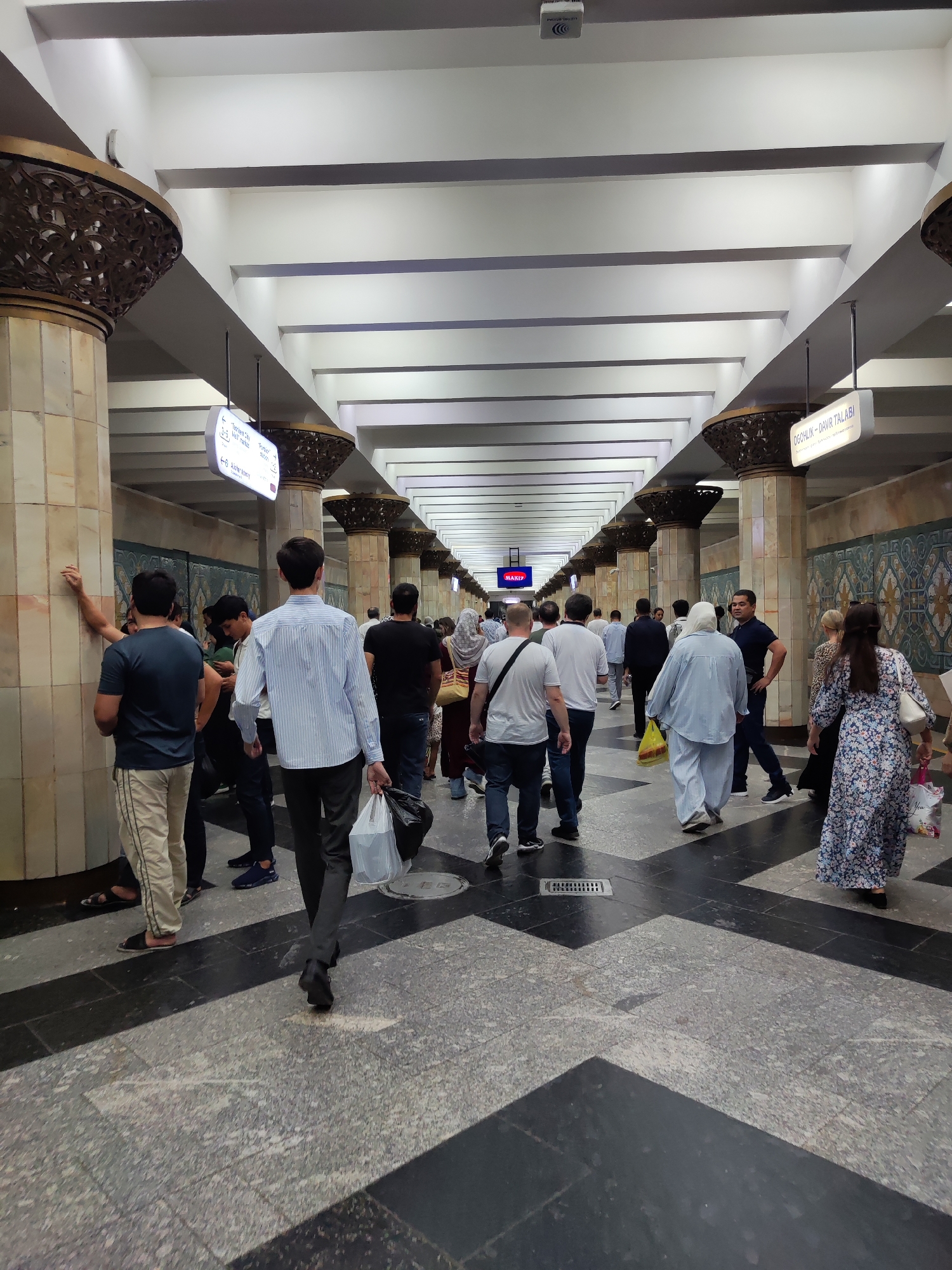 The image captures a scene within a bustling subway station in Tashkent, Uzbekistan, likely the Chorsu Metro station, known for its distinct architecture. A crowd of people is seen walking along a wide, ornate corridor. The ceiling features a series of angled white beams, and large, decorative pillars with intricate metallic filigree support the structure. The floor is a mosaic of dark and light grey tiles arranged in a geometric pattern.
Several signs are visible, some providing directional information with text in what appears to be Uzbek. One sign, hanging from the ceiling, points to "Toshkent City" and "Next Market". Another sign, further down, reads "QOG'HLIK - DAVR TALABI". The walls are decorated with patterned tiles in shades of blue and beige, and some sections have a textured appearance.
The people in the image are diverse in their attire and are engaged in the act of walking, presumably to catch a train or exit the station. Some individuals are carrying bags, including a person in the foreground with a transparent plastic bag and a black garbage bag. The overall atmosphere suggests a busy public transportation hub. The lighting is bright and even, illuminating the space effectively.
