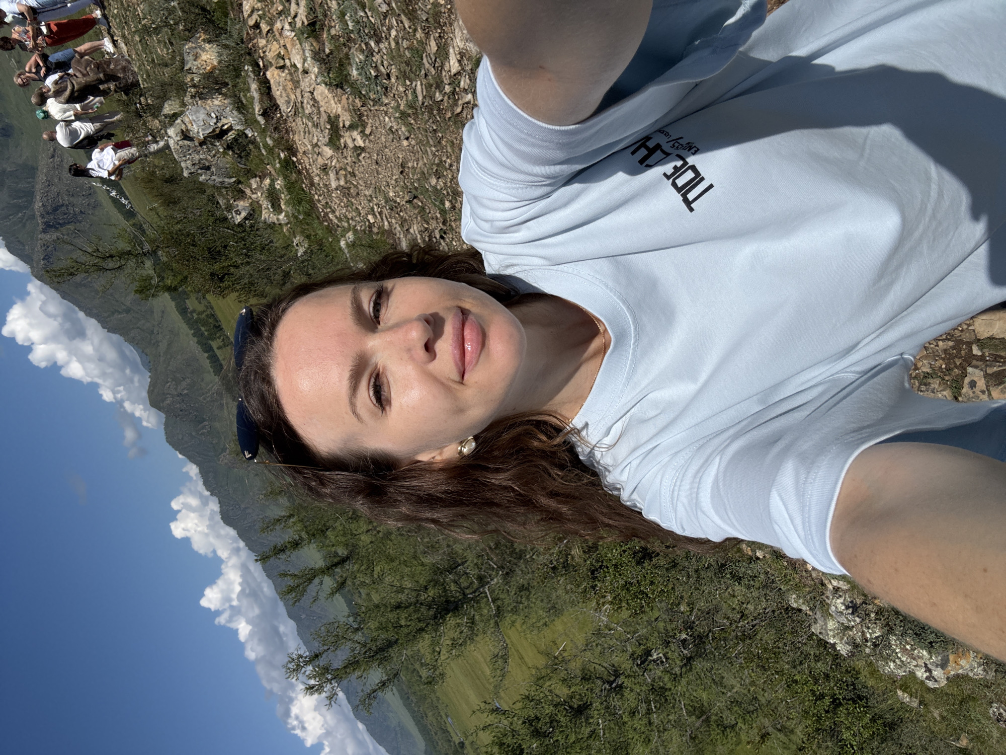 In a mountainous landscape in Bichiktu-Boom, Russia, a woman with long, wavy brown hair takes a selfie. She is wearing a light blue t-shirt with a black logo that reads "TIDE CH...". She has on large, light-colored earrings and sunglasses are perched on her head. In the background, a group of people are standing near a rocky embankment. The terrain is rugged with sparse vegetation and large rocks, with green valleys and steep, cloud-capped mountains in the distance. The sky is a clear blue with scattered white clouds.