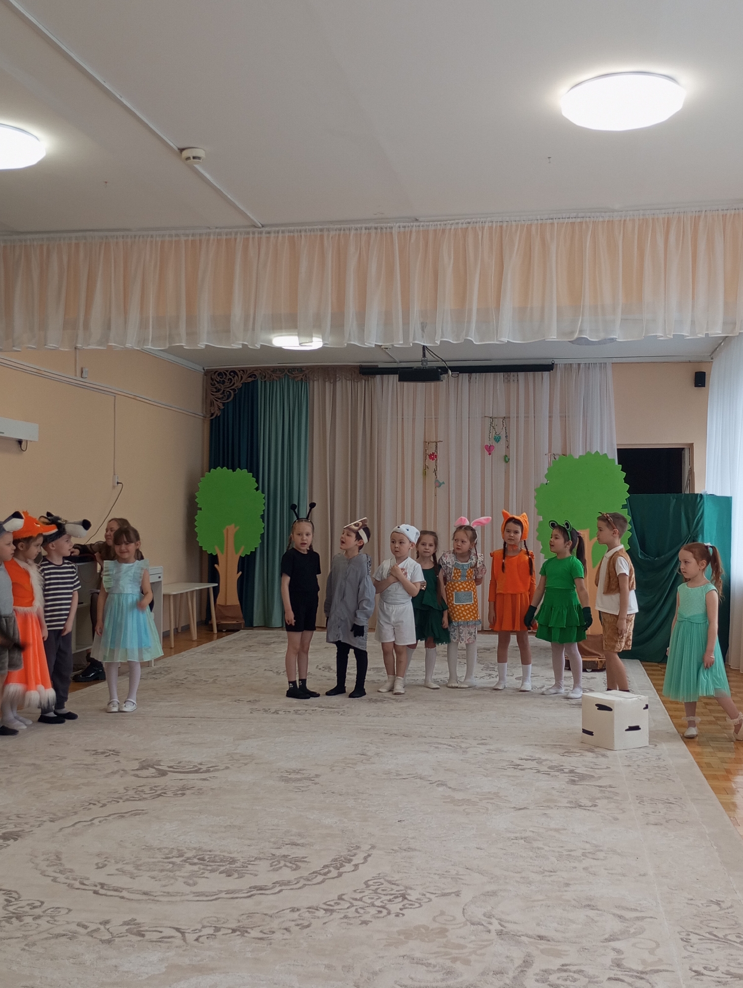 This full-shot image depicts a group of approximately 14 young children, likely aged between 4 and 8, engaged in a performance or rehearsal within an indoor hall, possibly a kindergarten or school in Izhevsk, Russia.
The children are dressed in a variety of animal-themed costumes, suggesting a nature or woodland play. On the far left, two children wear orange fox costumes with furry tails. Other children sport headbands with bunny ears, bear ears, or frog eyes, alongside full costumes like a green frog dress, an orange outfit, a light blue dress, or simpler attire like striped shirts and shorts. An adult is partially visible behind some children on the left.
The setting features light-colored walls and a high ceiling with modern circular LED lights and a visible smoke detector. Long, sheer cream-colored curtains adorn the upper walls, creating a decorative effect. The floor is covered with a large, light-toned rug featuring an elaborate, faded pattern. In the background, two green, cutout tree props define a stage area, behind which dark teal and beige curtains are drawn. A white cube prop with black markings is positioned on the rug near the front right.
The children are arranged in lines, mostly facing towards the right side of the frame, appearing attentive and focused on the activity. The bright lighting suggests it is daytime. The overall atmosphere is cheerful and organized, typical of a children's event. No visible text is legible in the image.