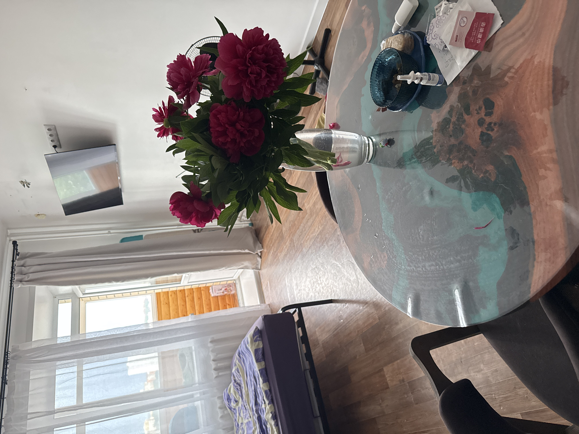 The image shows a sparsely furnished room, likely an apartment. In the foreground is a round table with a swirled, possibly resin-coated surface. On the table are various items including what appears to be a small blue dish, medication, and a vase of deep red peonies. The room has a window covered by sheer white curtains, allowing some natural light. A bed or sofa with a purple blanket is visible near the window. A flat-screen television is mounted on the wall, which is painted white. The floor is wood-grain. The overall impression is a simple, somewhat cluttered, domestic scene. There is nothing in the image to indicate it is in Perm, Russia.