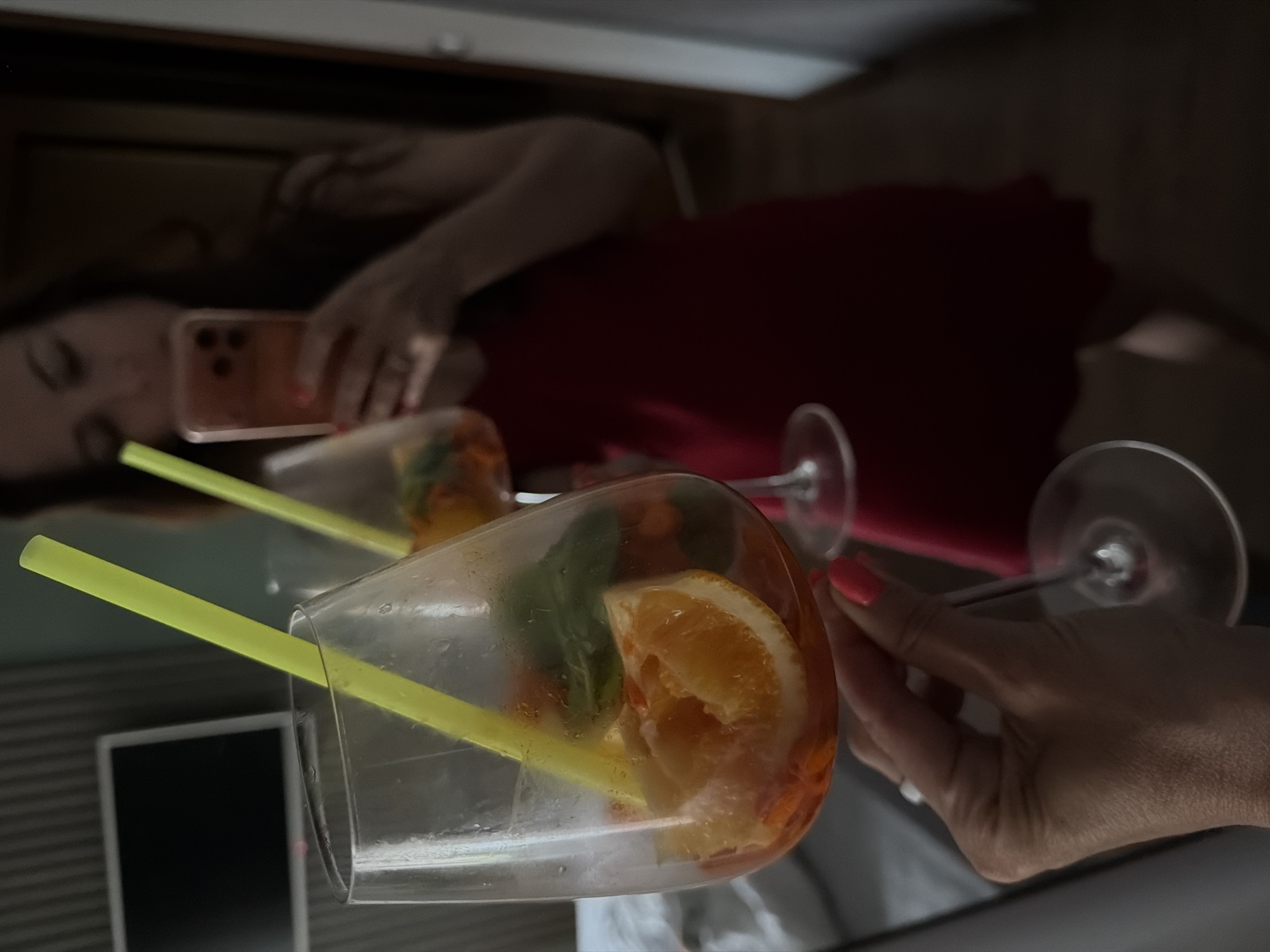 The scene is situated in Birobidzhan, Russia. The image captures a mirror selfie featuring a person holding a large stem glass containing an orange-hued beverage. The drink is garnished with an orange slice, a green leaf (likely mint or basil), and a bright yellow plastic straw. The person's hand holding the glass exhibits pink painted fingernails and a ring on one finger. The reflection shows the individual wearing a red garment, possibly a dress or top, and holding a light-colored smartphone with a visible multi-lens camera array to capture the photograph. Their face is out of focus. A second identical drink, also garnished with an orange slice and yellow straw, is partially visible in the reflection to the right. In the background of the reflection, a wall-mounted flat-screen television with a dark screen is visible against a subtly textured or slatted wall. To the left, a soft, light-colored surface, possibly bedding from a bed or sofa, is present. The overall environment suggests an indoor setting, likely a hotel room or apartment, with subdued lighting.