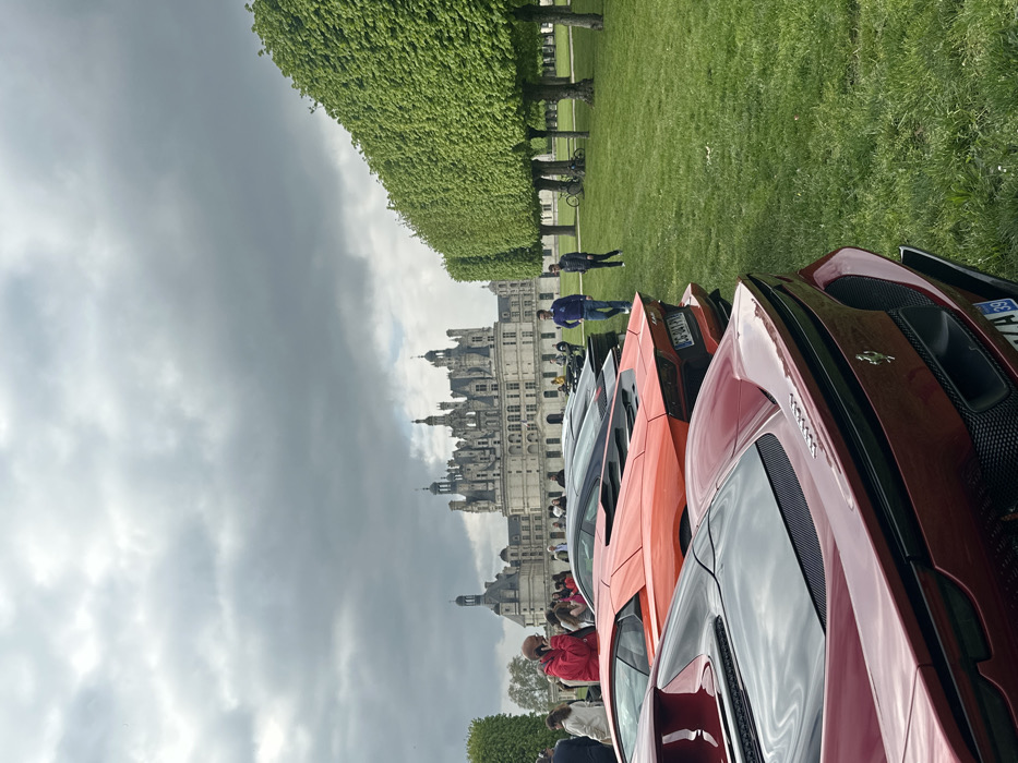 The image shows a red Ferrari parked in front of a bright orange Lamborghini. The Lamborghini is parked in front of a black car. The cars are parked in a grassy field. The Chateau de Chambord, a large castle in the Loire Valley, is visible in the background. The castle is made of white stone and has many towers and turrets. It is surrounded by a moat. The sky is cloudy. There are a few people walking around. The coordinates provided, 47.615 latitude and 1.519 longitude, place the scene in the Chateau de Chambord's grounds in the city of Chambord, France.