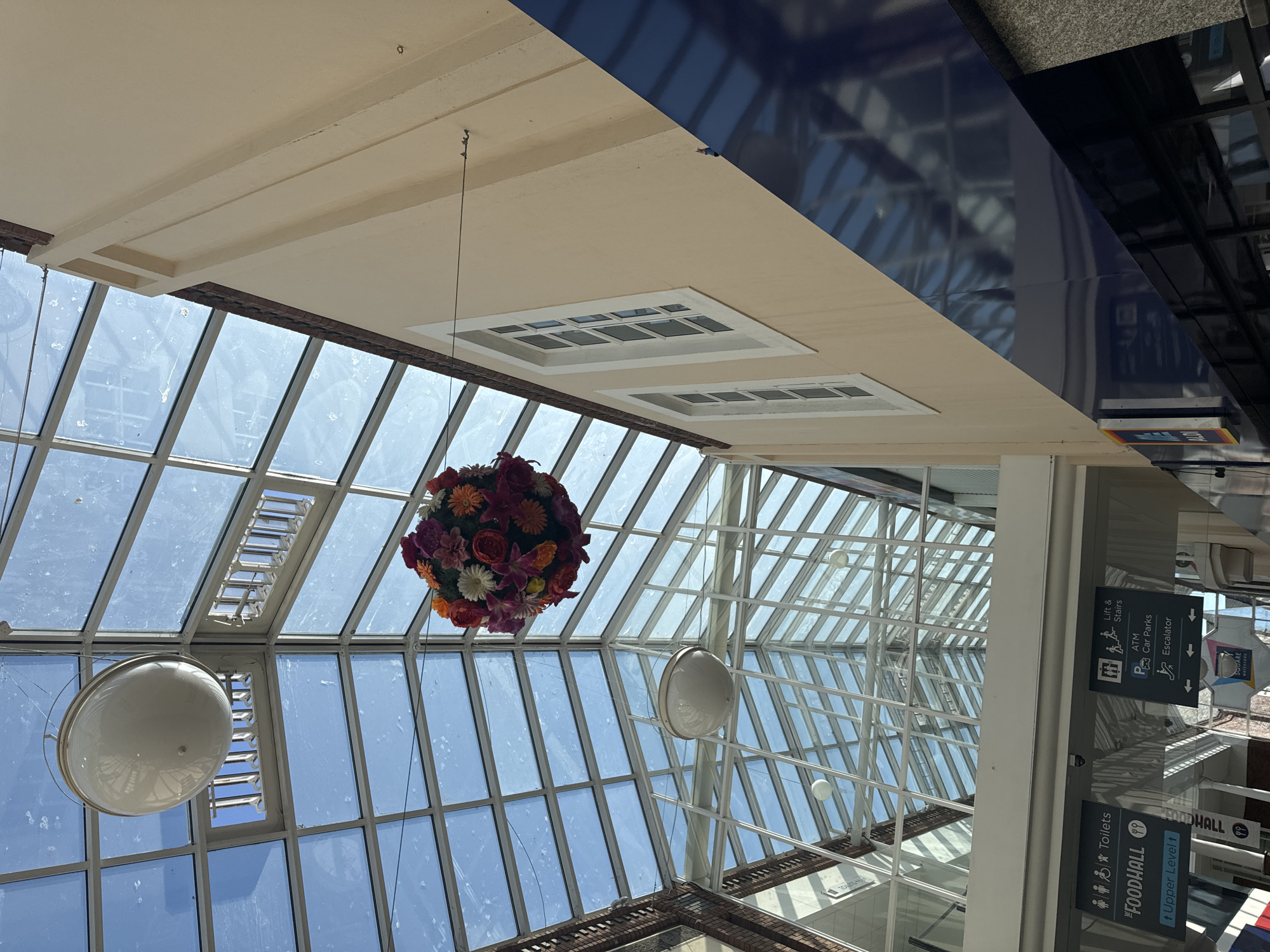 This image shows the interior of a modern shopping center, likely a covered walkway or atrium, with a prominent glass ceiling. The ceiling is divided into a grid of large panes, allowing natural light to flood the space. Suspended from the ceiling are two large, circular white light fixtures and a colorful spherical floral arrangement.
The walls of the building are painted in a light beige, with some sections featuring exposed brickwork. There are several rectangular windows with white frames on the beige walls. The floor is not clearly visible, but a blue reflective surface runs along the bottom right of the image, suggesting shop fronts or displays.
Below the glass ceiling, directional signage is visible. One sign indicates "Toilets" and "FOODHALL" with an arrow pointing to "Upper Level". Another sign lists "Lift & Stairs", "ATM", "Car Parks", and "Escalator".
The architecture and signage suggest this is a public commercial space. Based on the signage "Waterford" on a t-shirt visible at the bottom of the frame, and the overall style, this location is likely the **Market Cross Shopping Centre in Waterford, Ireland**.