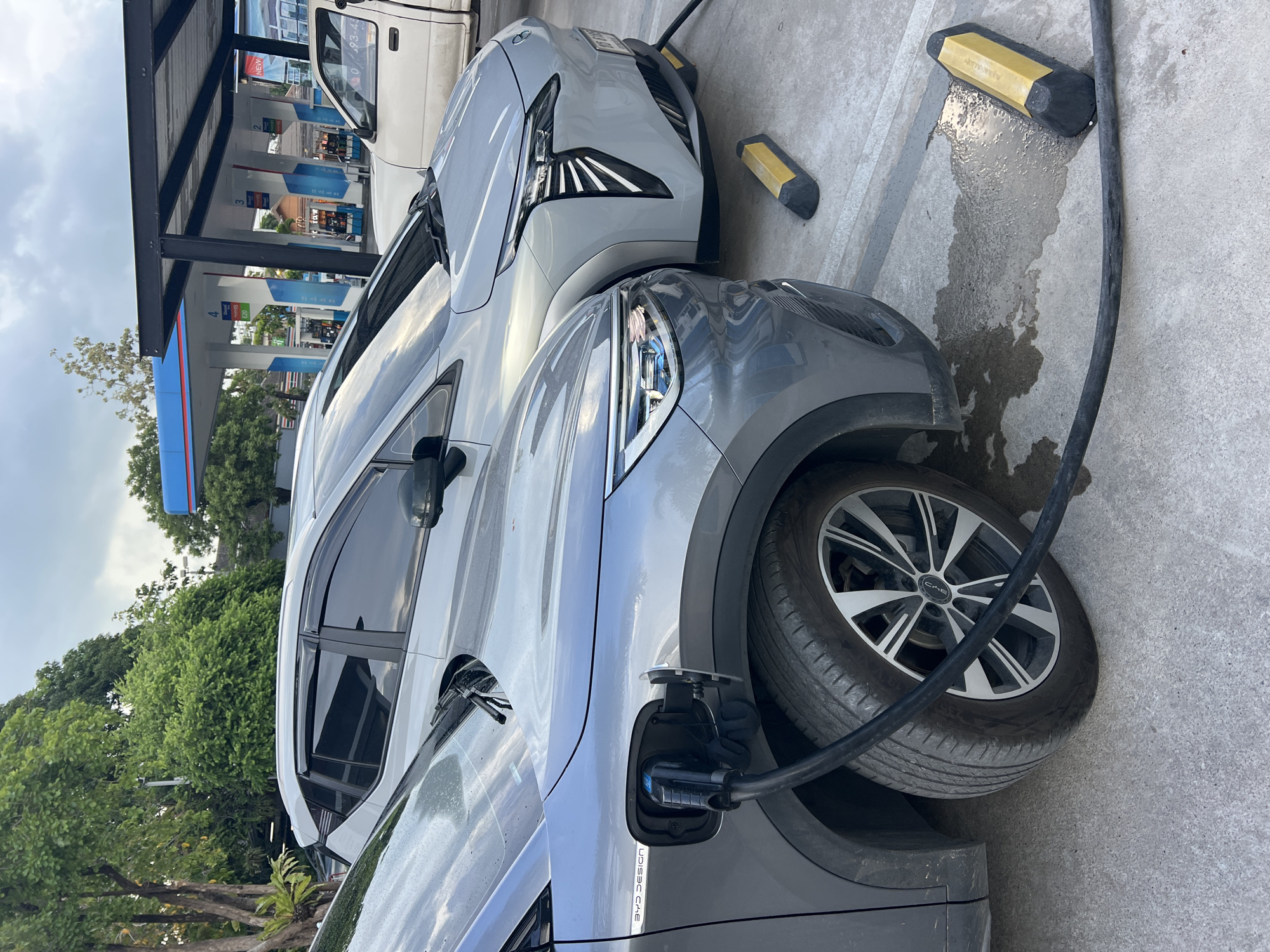 In Baan Bo Phut, Thailand, two electric vehicles are being charged at a charging station. The gray vehicle in the foreground has a charging cable connected to its charging port. The charging station is located under a gas station canopy. A white vehicle is also parked nearby. The surrounding environment includes trees and the concrete parking surface.