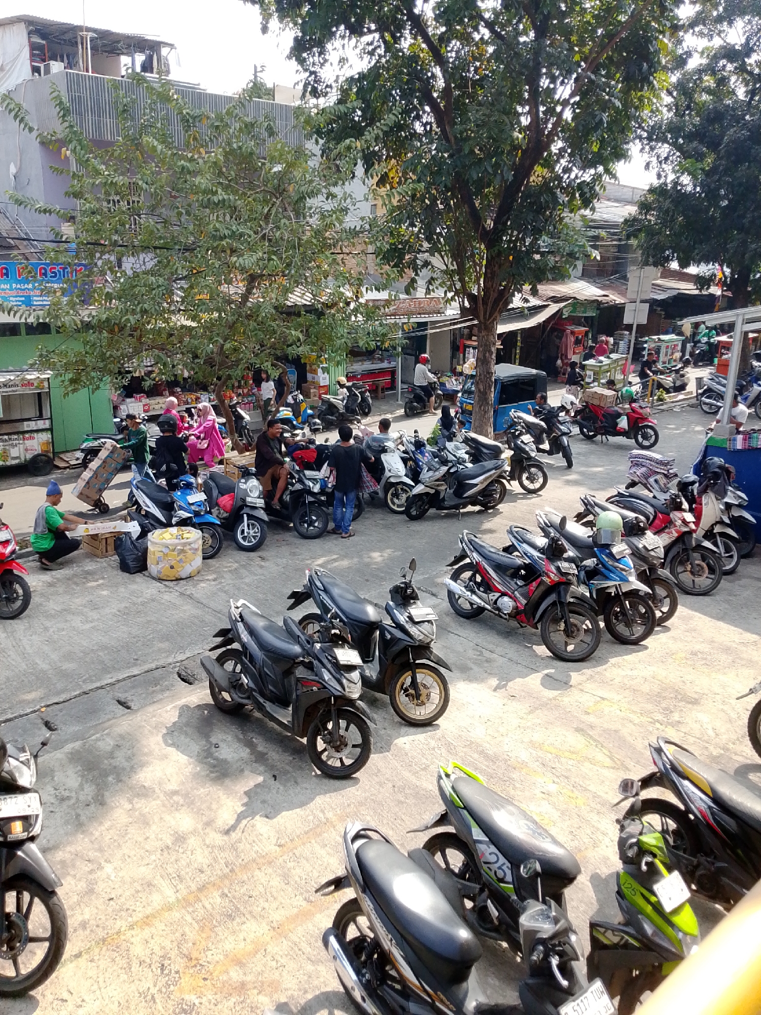 The image depicts a street scene in the city of East Jakarta, Indonesia. A large number of motorcycles are parked on a paved surface, suggesting a busy marketplace or public area. Several trees with dense foliage provide shade. In the background, buildings with various signs and awnings are visible, indicating commercial activity. Some people are seen walking or standing near the motorcycles and stalls. The overall impression is a bustling, everyday scene in an Indonesian urban environment.