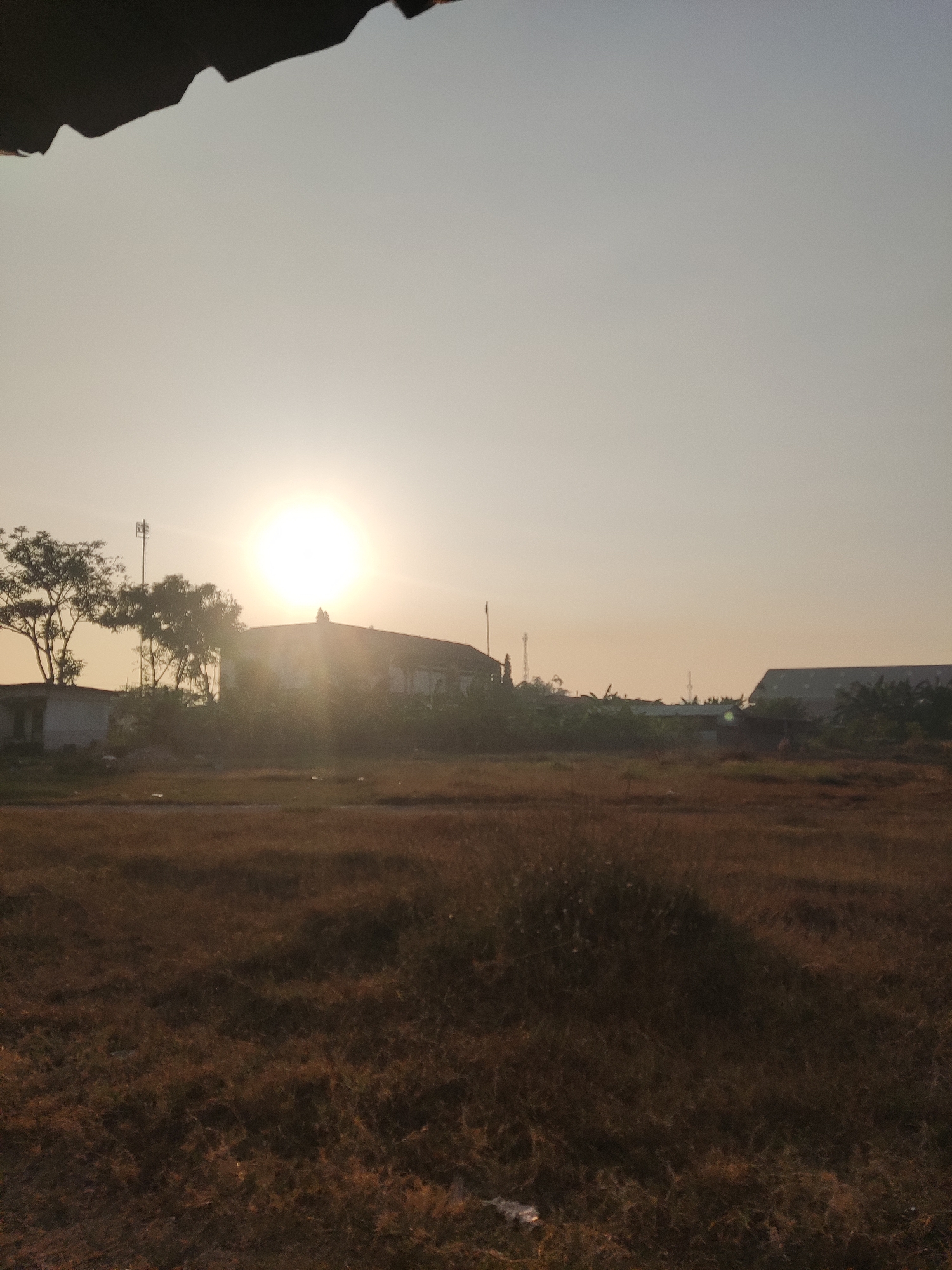 A photograph captures a scene in Banjarkemantren, Indonesia, during sunrise. The sun, a bright orb, is positioned in the upper left quadrant of the image, casting a warm glow across the sky, which graduates from a pale yellow near the horizon to a light gray at the top. In the mid-ground, a building with a dark, angled roof is visible, silhouetted against the sun. To the left of the building, a solitary telecommunications tower stands tall. A cluster of trees with sparse foliage is situated to the left of the tower and building. The foreground consists of dry, golden-brown grass, indicative of a rural or undeveloped area. The overall impression is of a quiet, possibly industrial or semi-urban fringe, as suggested by the presence of buildings and the tower.