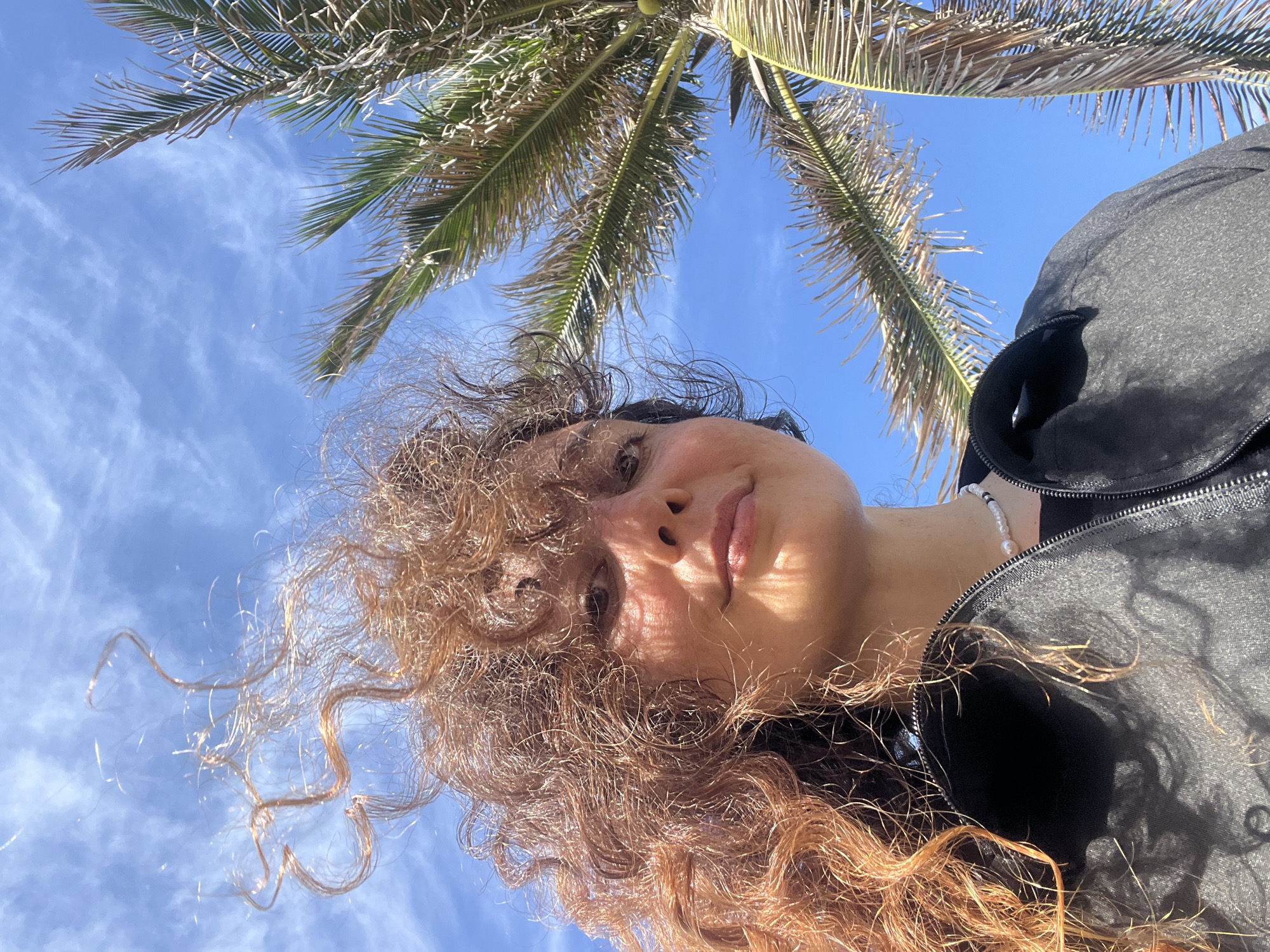 A low-angle, wide selfie is captured outdoors under a bright, clear blue sky with wispy white clouds. A woman with curly, reddish-brown hair is positioned in the left foreground, her face angled upwards and slightly to the right, with a soft smile. She is wearing a dark jacket and a pearl necklace. Her hair is blowing in the wind. To the right of the woman, a portion of a palm tree with green and brown fronds is visible against the sky. The scene suggests a sunny, breezy day, likely in a tropical or subtropical location. The image does not provide enough information to determine the specific location within Phu Thuy Ward, Vietnam.